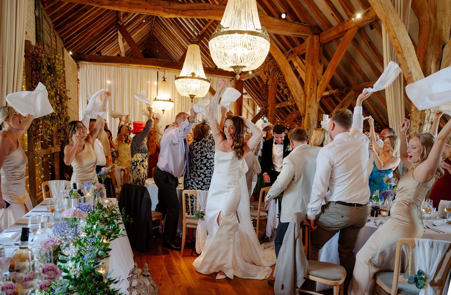 Elegant wedding reception venue with crystal chandeliers and rustic wooden beams decorated in white linens and floral accents.