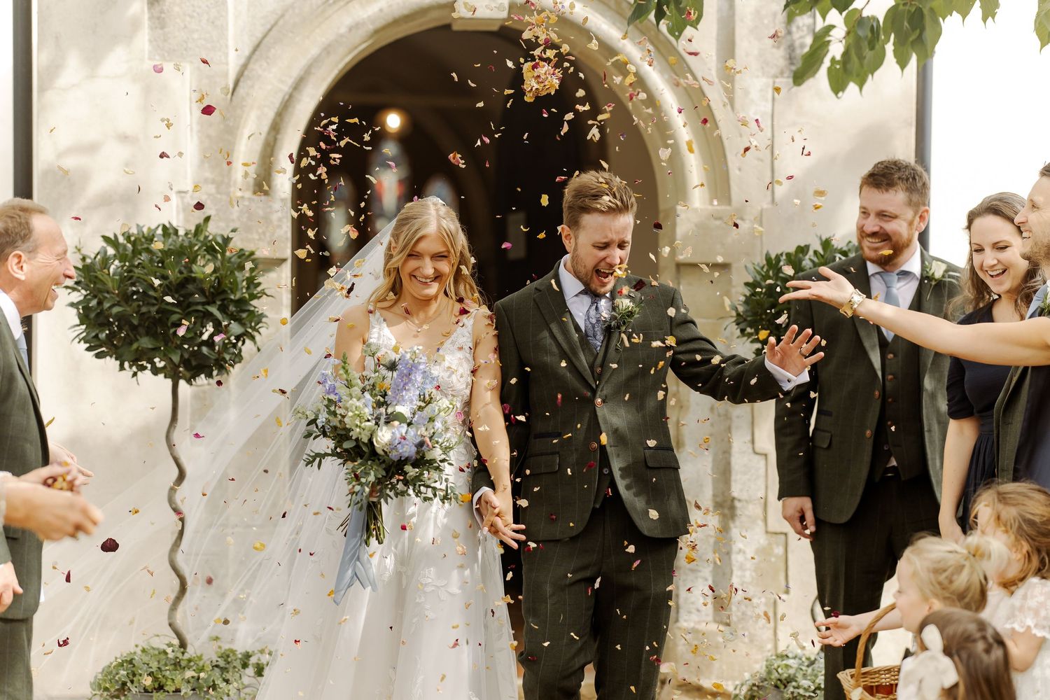 Bride and groom exit church amid confetti shower, guests celebrating joyful outdoor wedding ceremony.