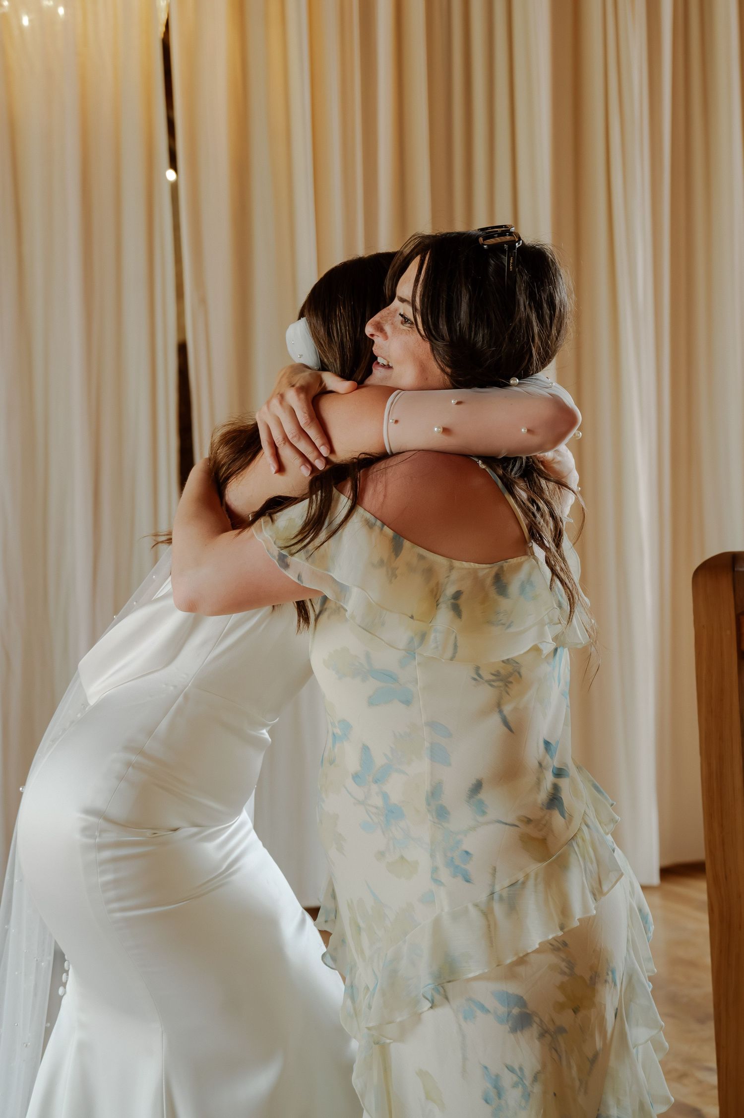 Two people in white and floral dresses embrace in an emotional moment against a cream curtain backdrop.