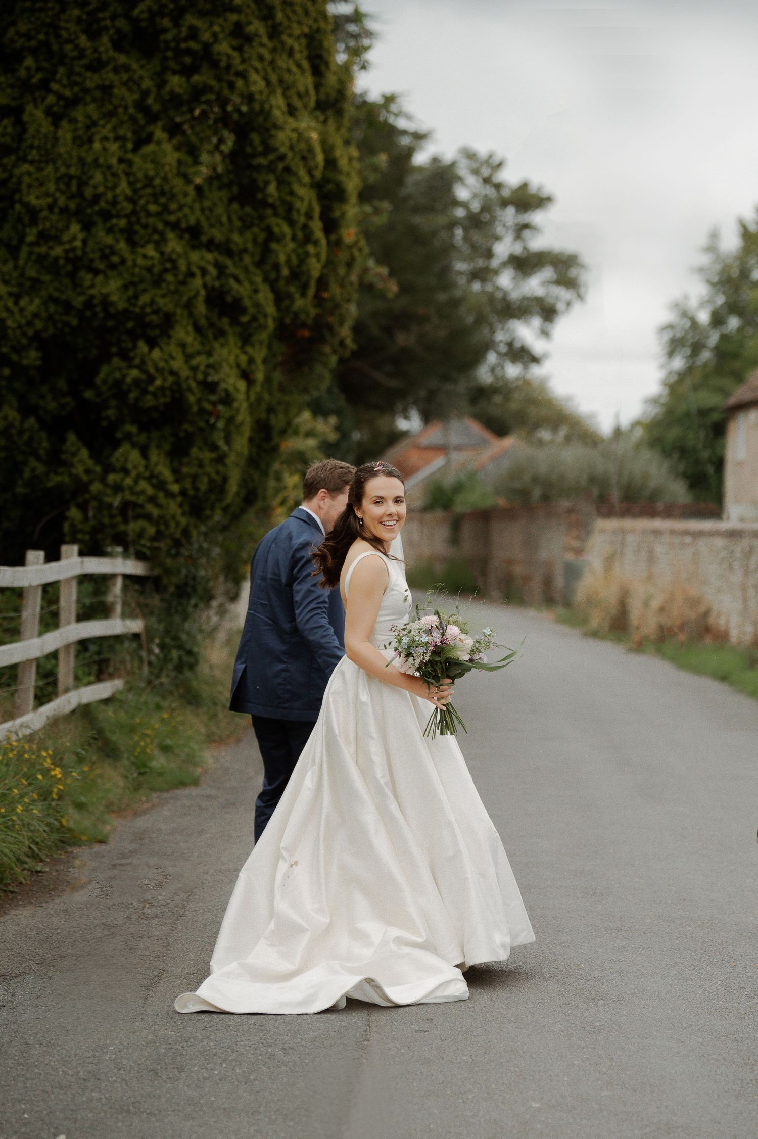 A couple in wedding attire share intimate moments along a countryside path with white fencing and greenery.