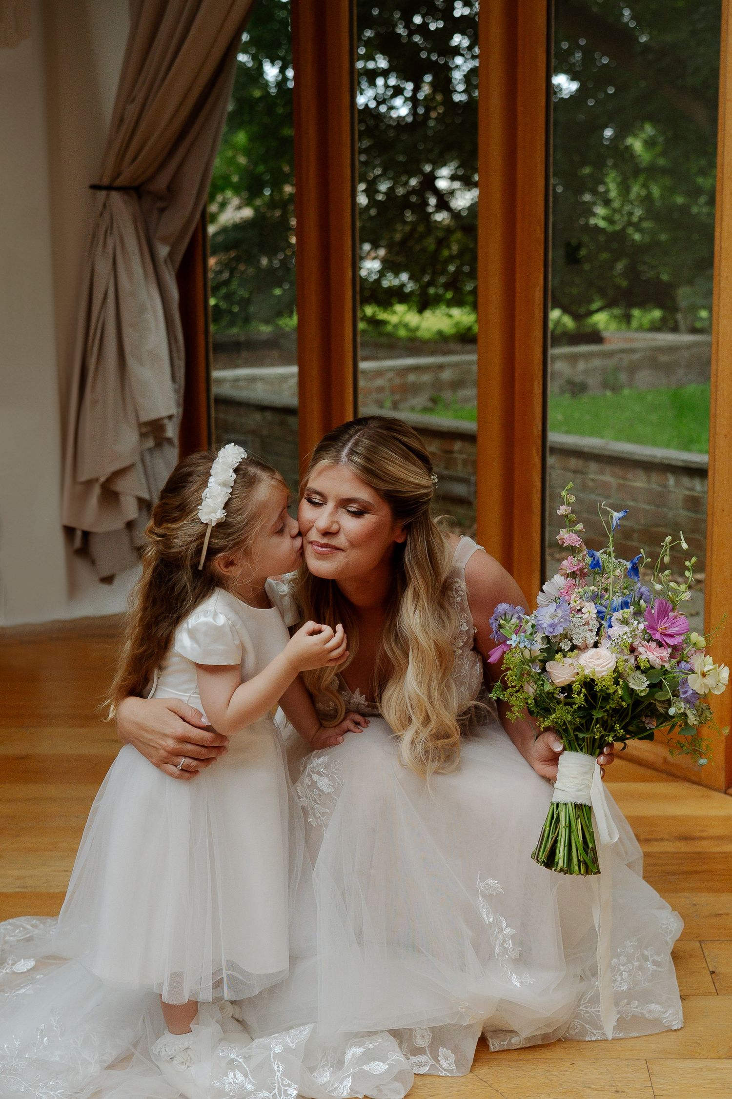 Two people in white dresses share a tender moment by a window with colorful flowers and a bouquet.