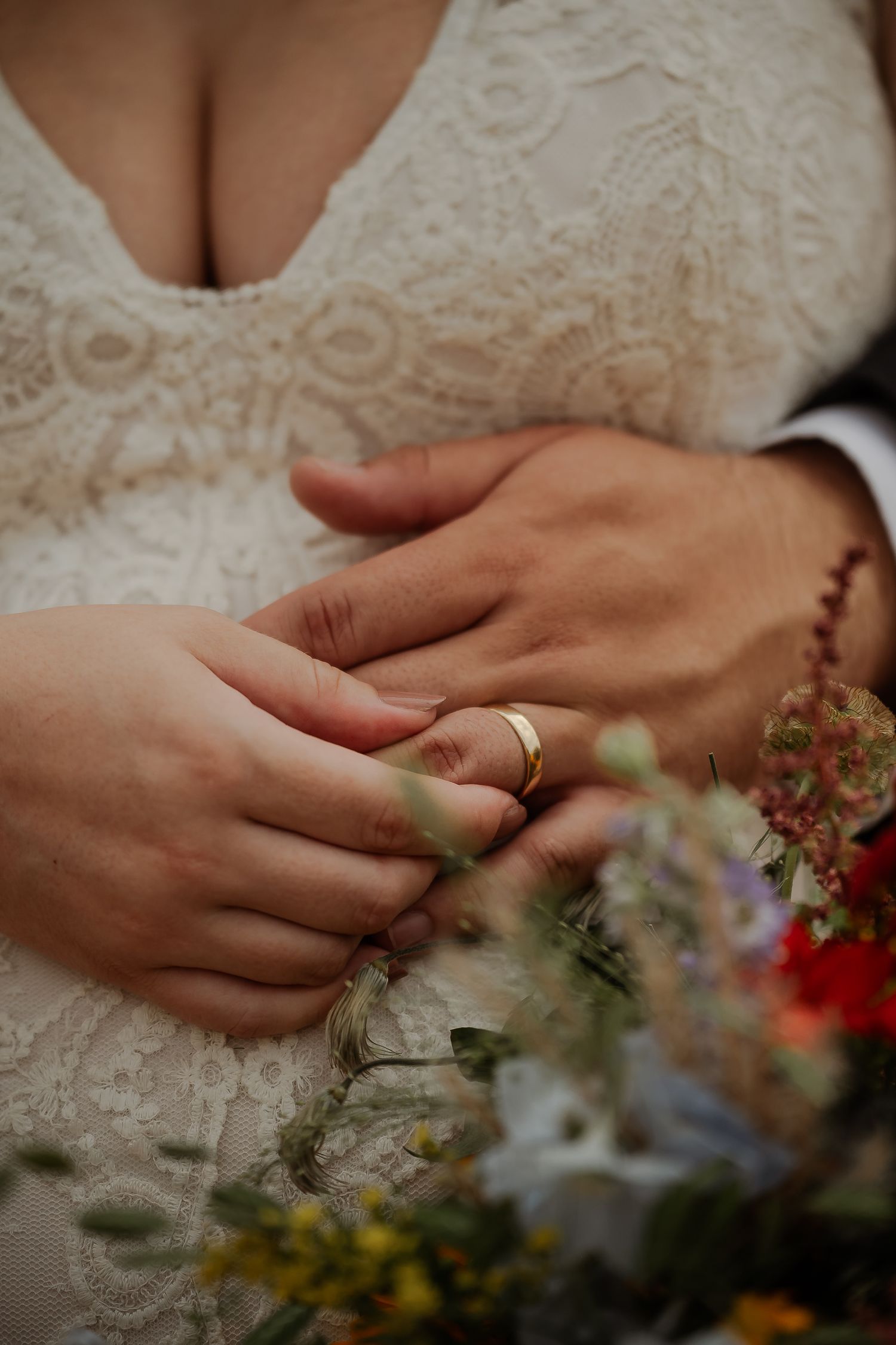 Close-up of newlyweds hands with gold wedding ring, bride in lace dress holding colorful wildflower bouquet.
