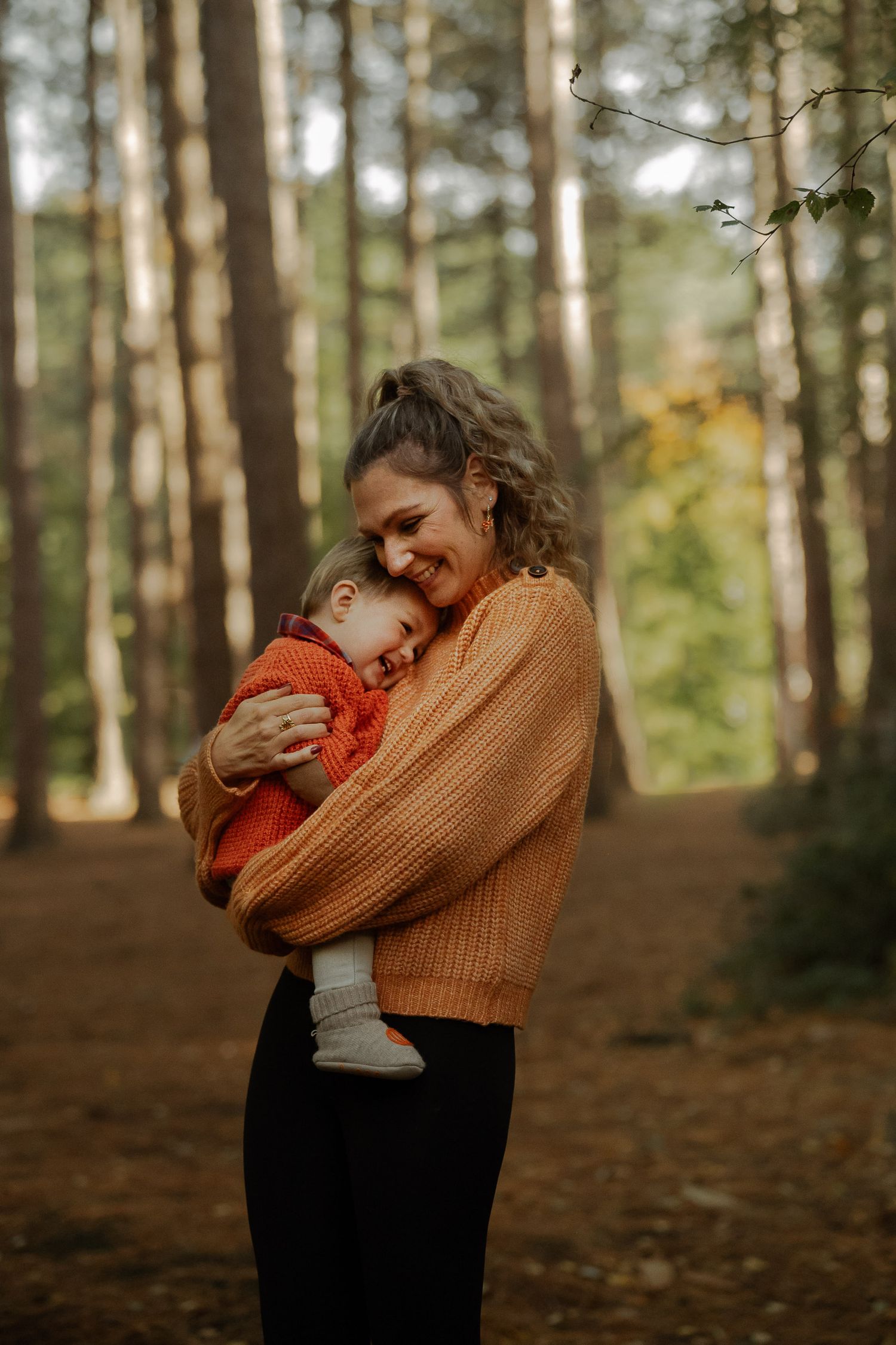 A smiling woman in an orange sweater tenderly holds a sleeping baby in an autumn forest setting.