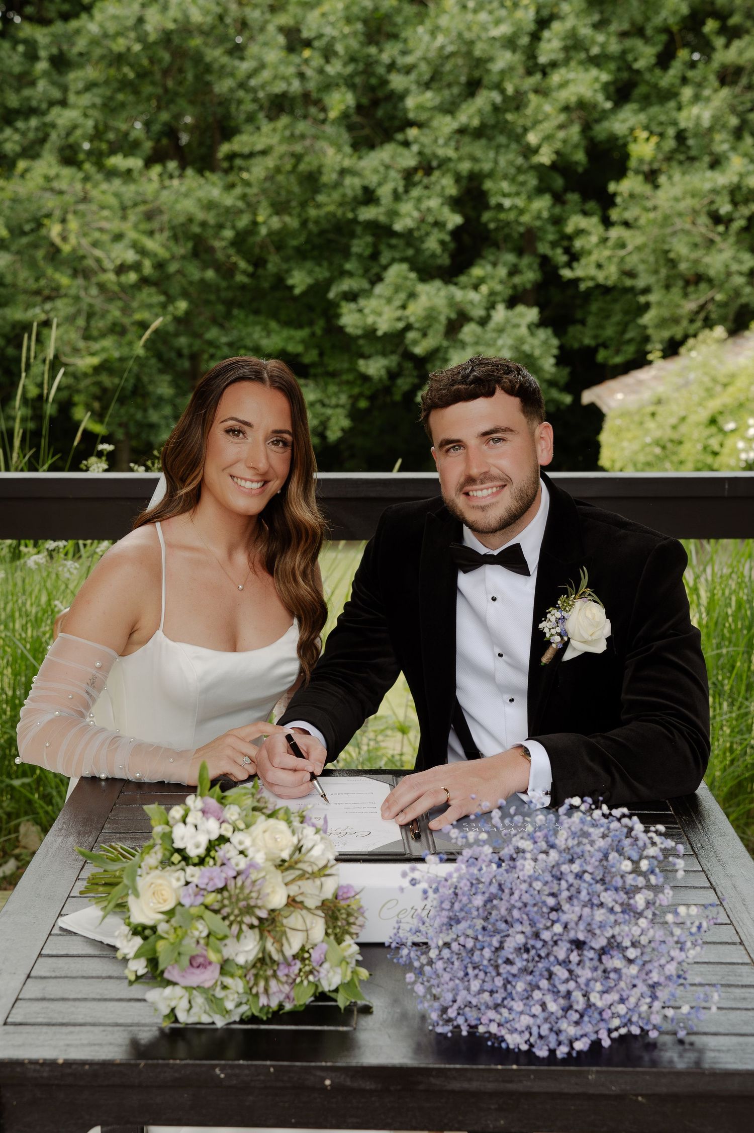 A couple sits at a rustic table with purple hydrangea and white floral bouquets during their wedding celebration.
