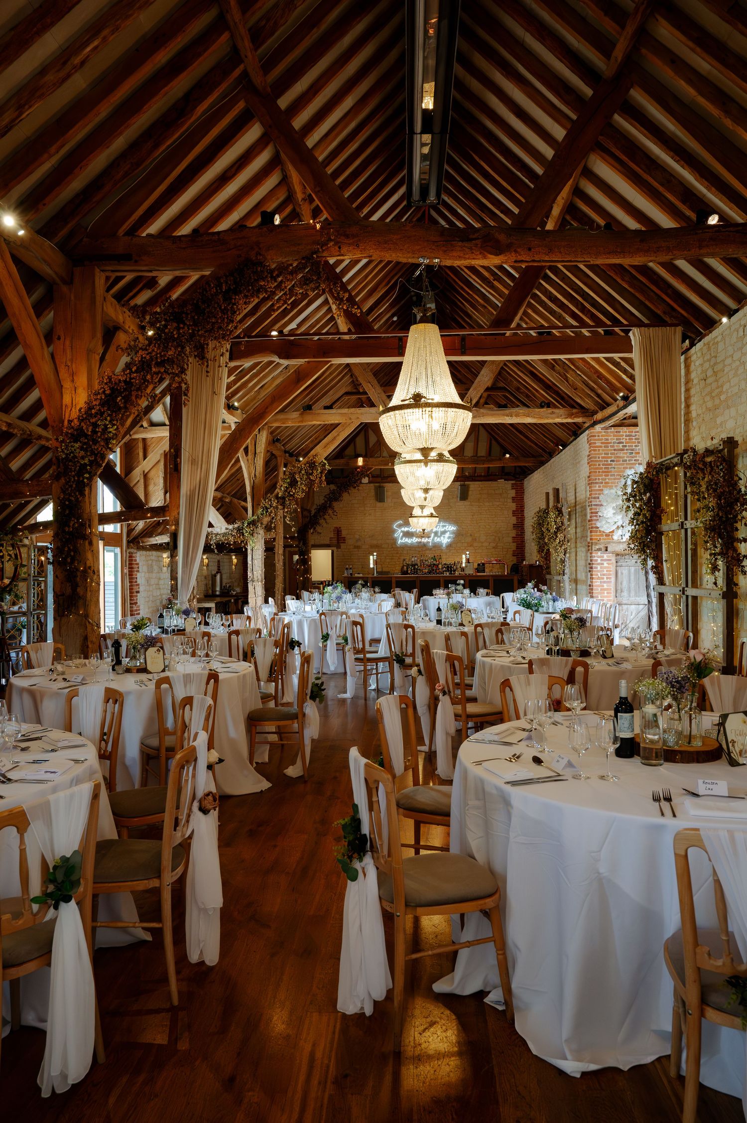Elegant rustic barn wedding reception venue with white tablecloths and chandeliers hanging from wooden beams.