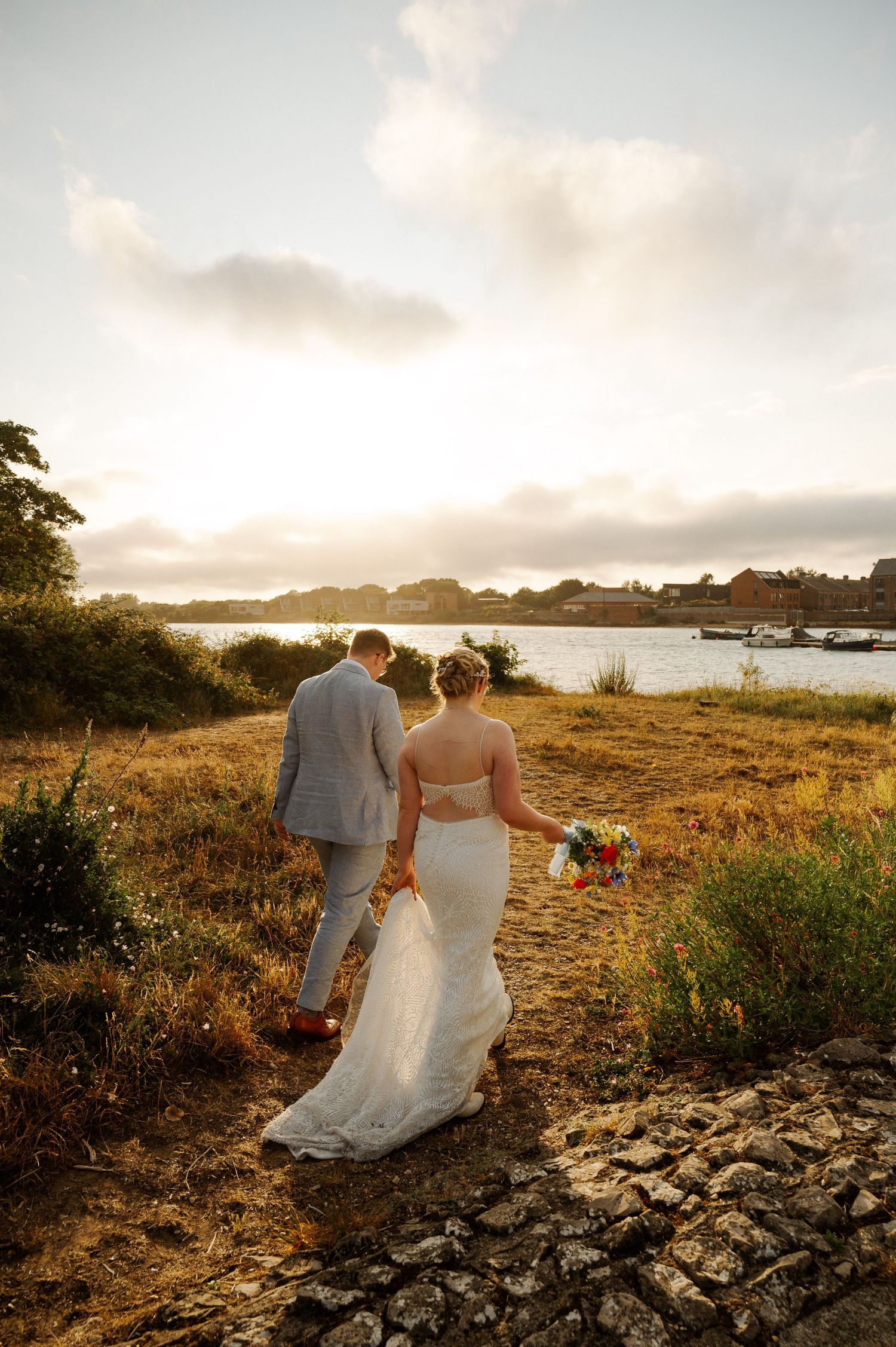 A couple in wedding attire walks along a scenic coastal path during golden hour at sunset.
