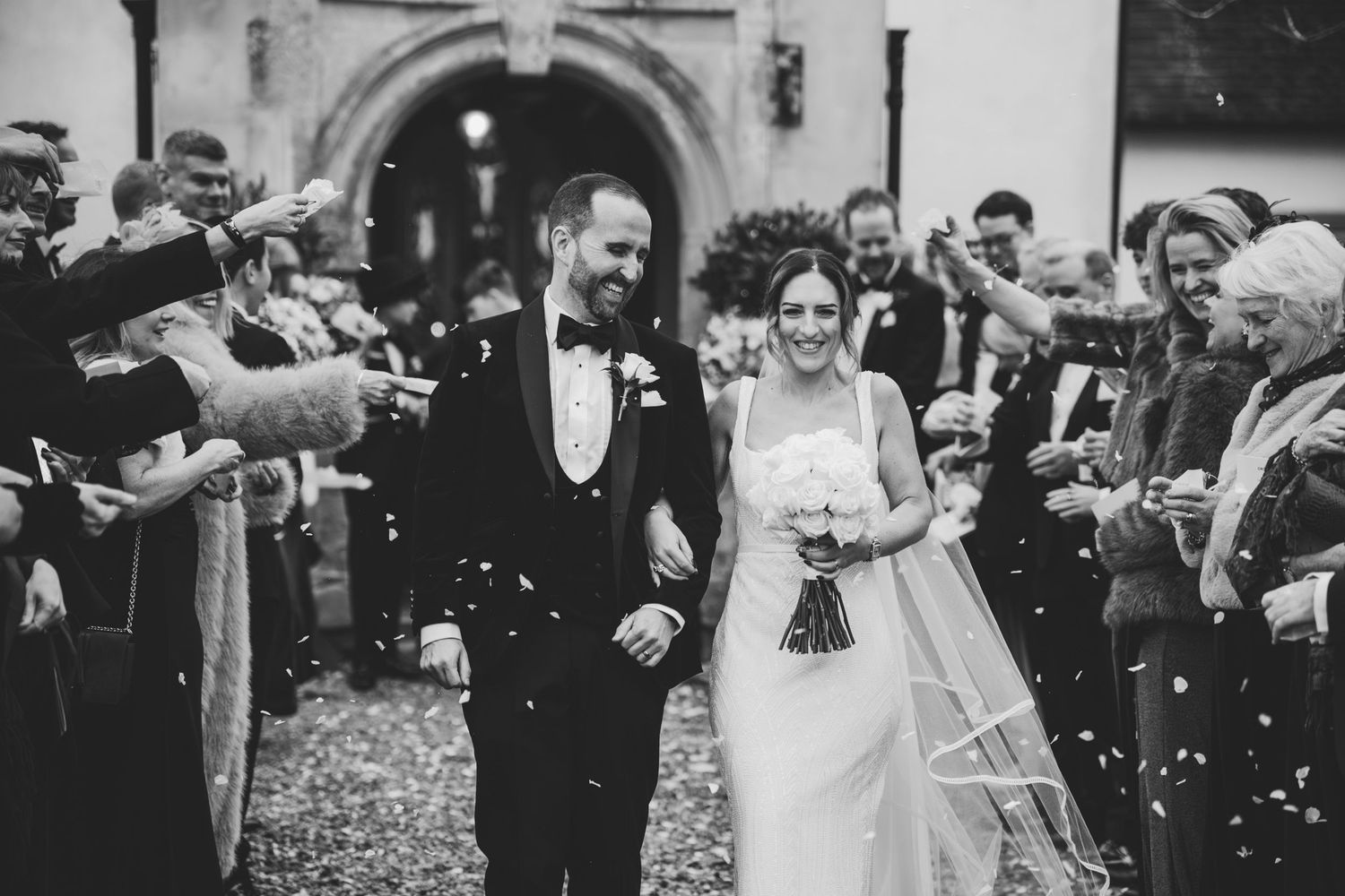 Wedding couple exits church ceremony while guests throw confetti in joyful black and white celebration moment.