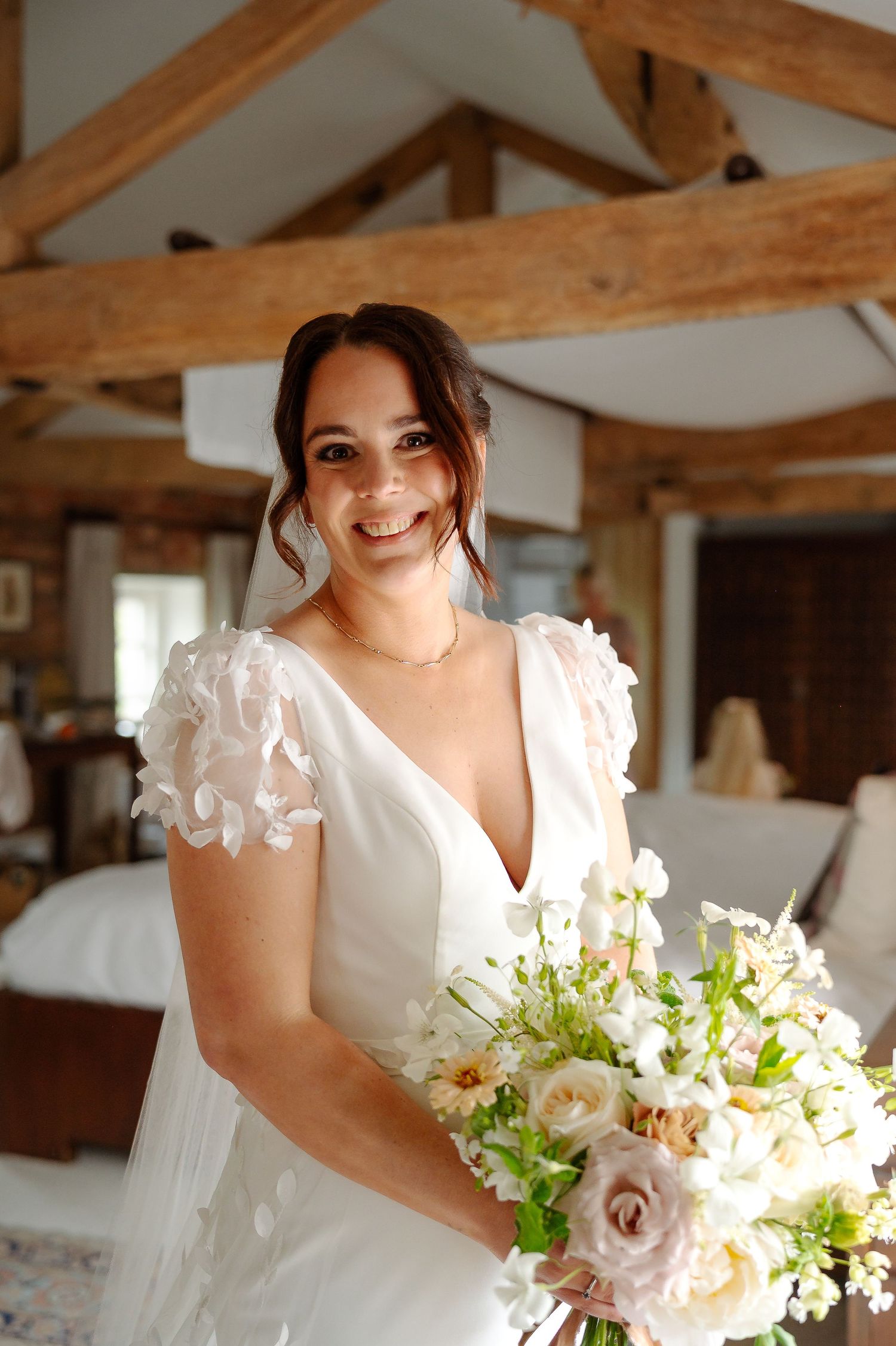 Smiling bride in white floral gown holding elegant bouquet of white and blush roses in rustic barn venue.