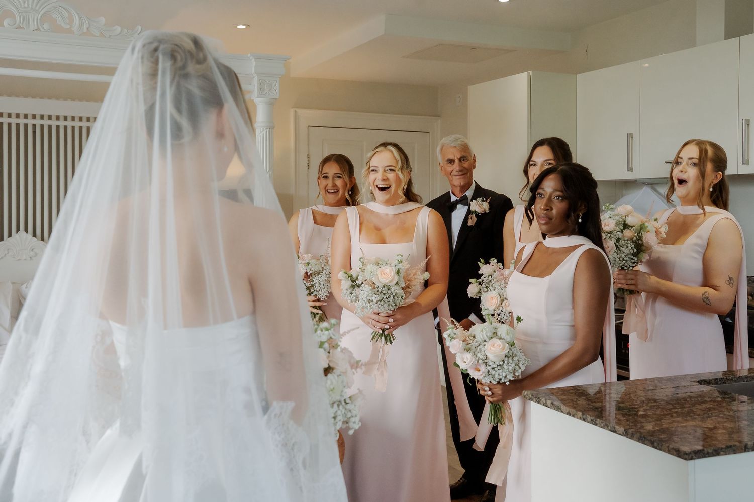 Bridesmaids react with joy and surprise seeing bride in wedding dress and veil during emotional first look moment.