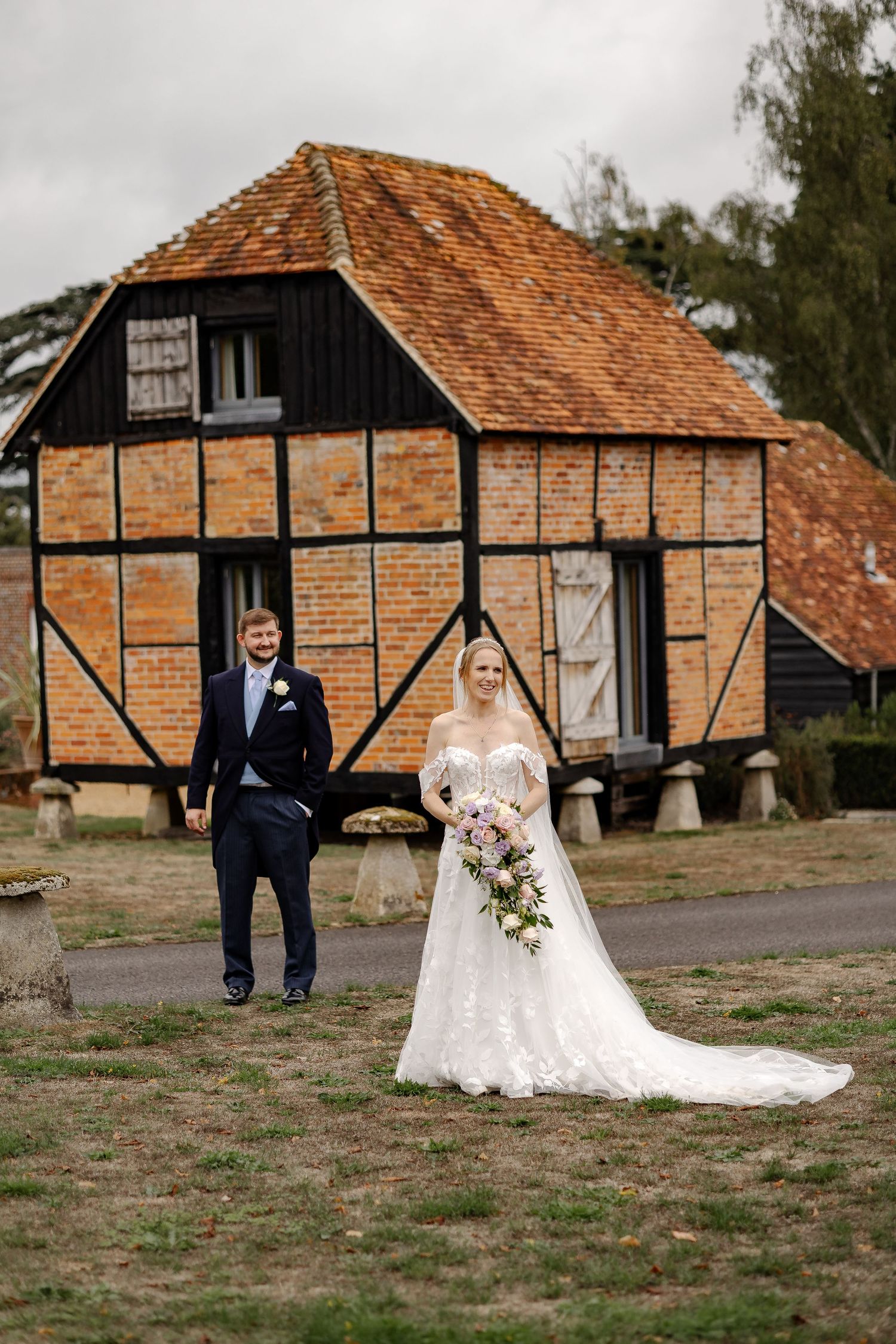 Bride in lace gown holding bouquet stands with groom in navy suit before rustic brick and timber barn wedding venue.