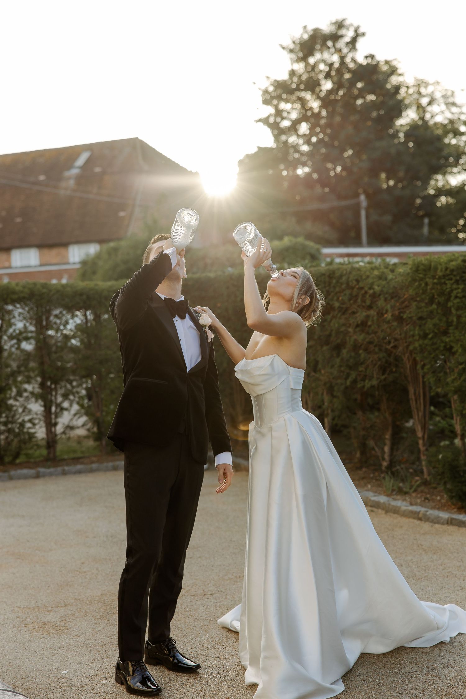 Bride and groom toasting champagne glasses in golden sunset light, she in strapless white gown, he in black tuxedo.