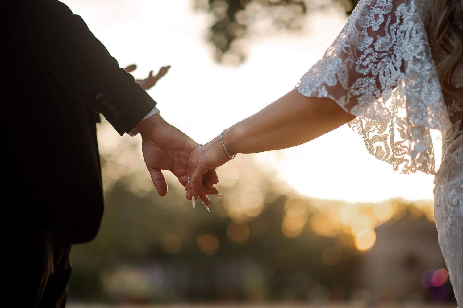 Bride and groom holding hands at sunset, wedding couple closeup with golden bokeh background.