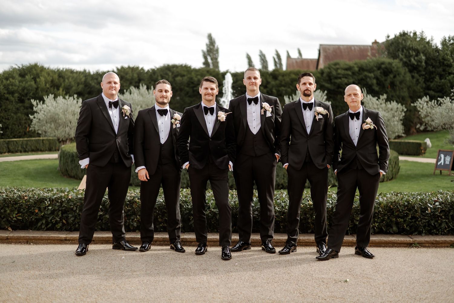 Six groomsmen in black tuxedos with bow ties pose outdoors at a wedding venue with manicured gardens.