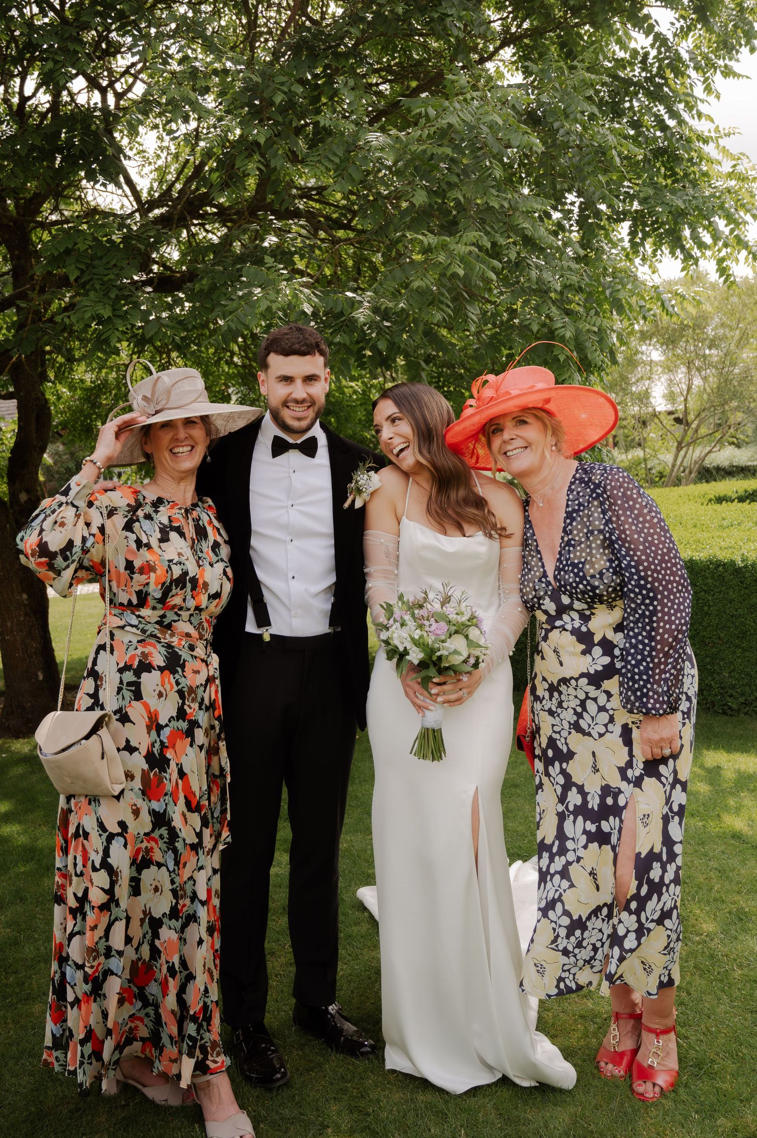 Wedding party poses outdoors in formal attire with floral dresses and bouquets under green trees.