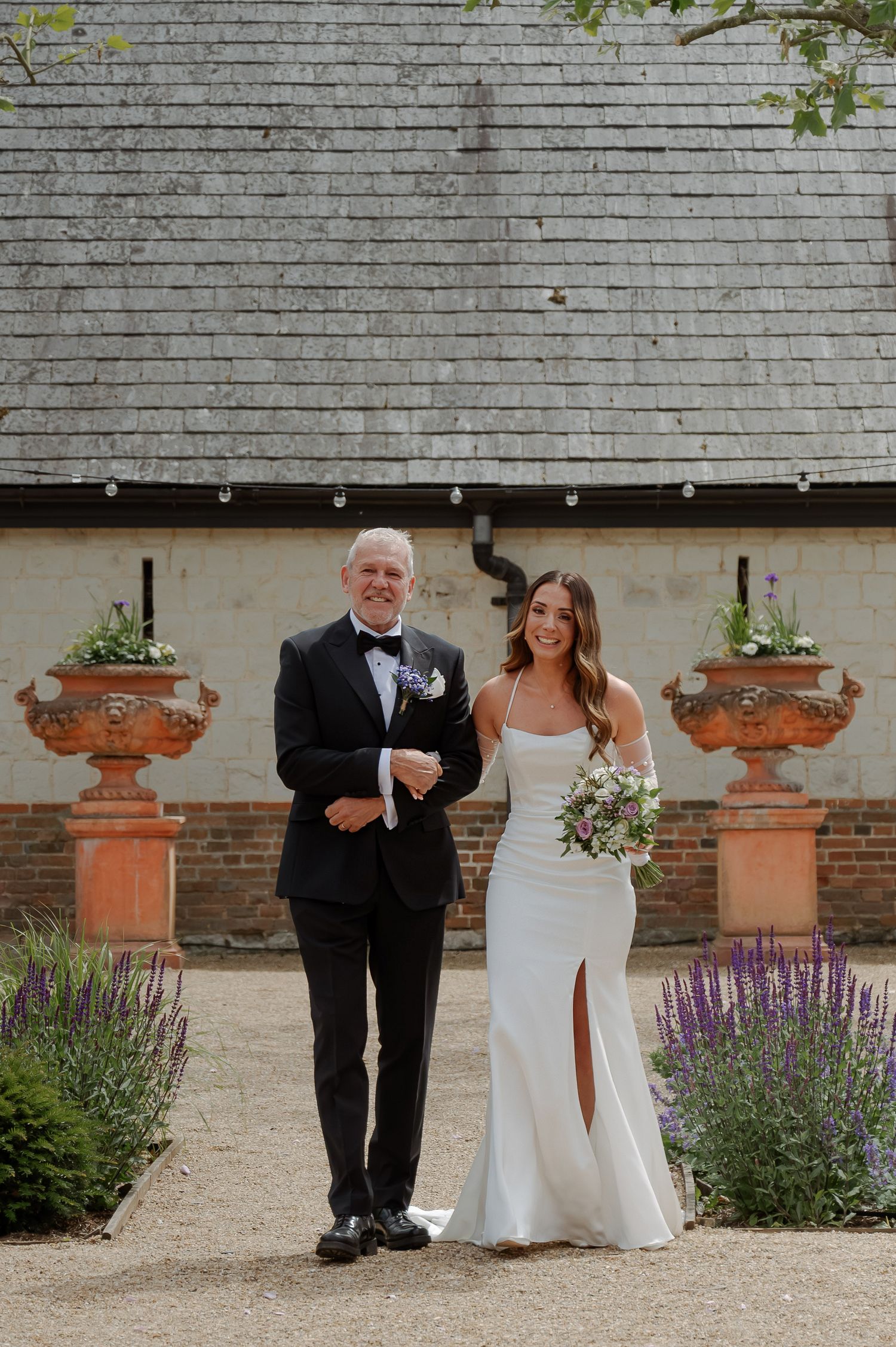 Wedding couple poses in a garden setting with terracotta urns and purple flowers.