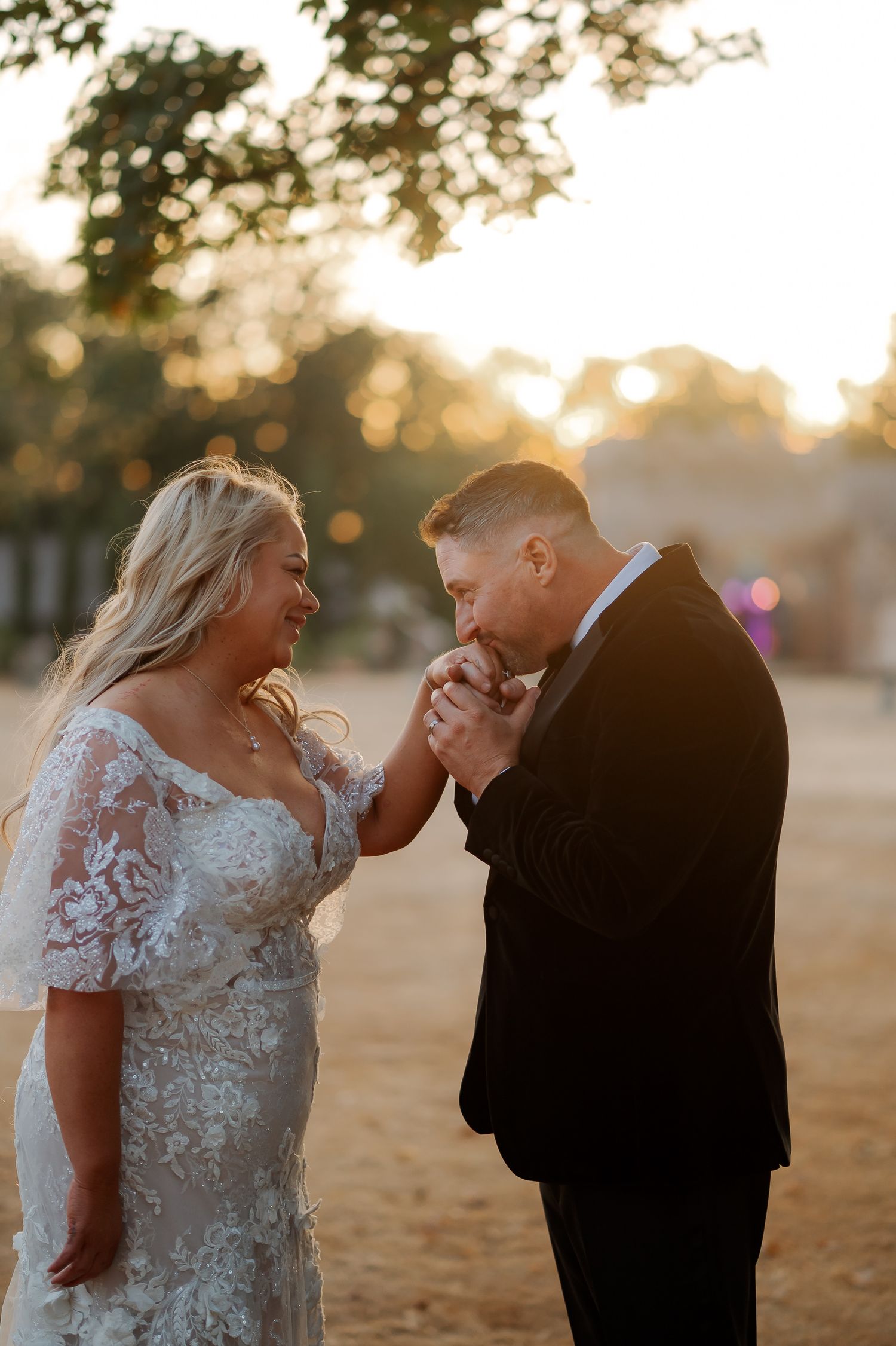 Groom kissing bride's hand at golden hour sunset, bride wearing lace flutter sleeve wedding dress outdoors.