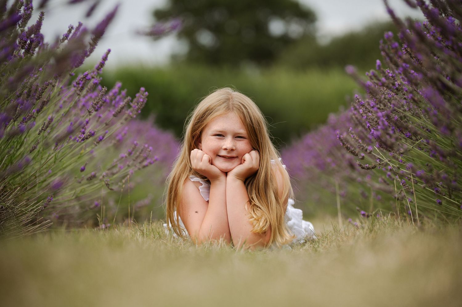 Young girl with long blonde hair smiling in a lavender field, resting chin on hands among purple blooms.