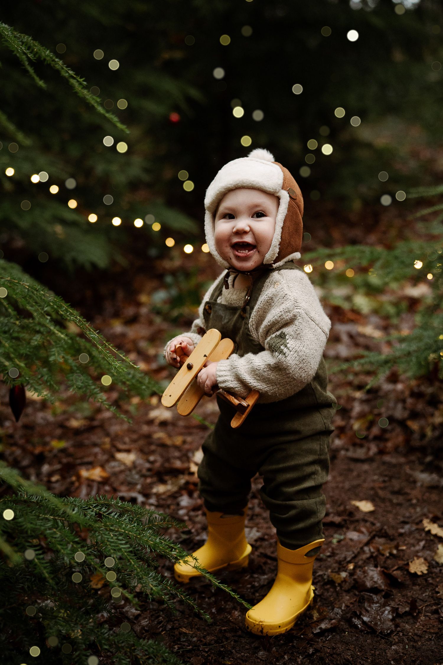 Smiling toddler in yellow boots, aviator hat, holding wooden toy plane near Christmas tree with bokeh lights.