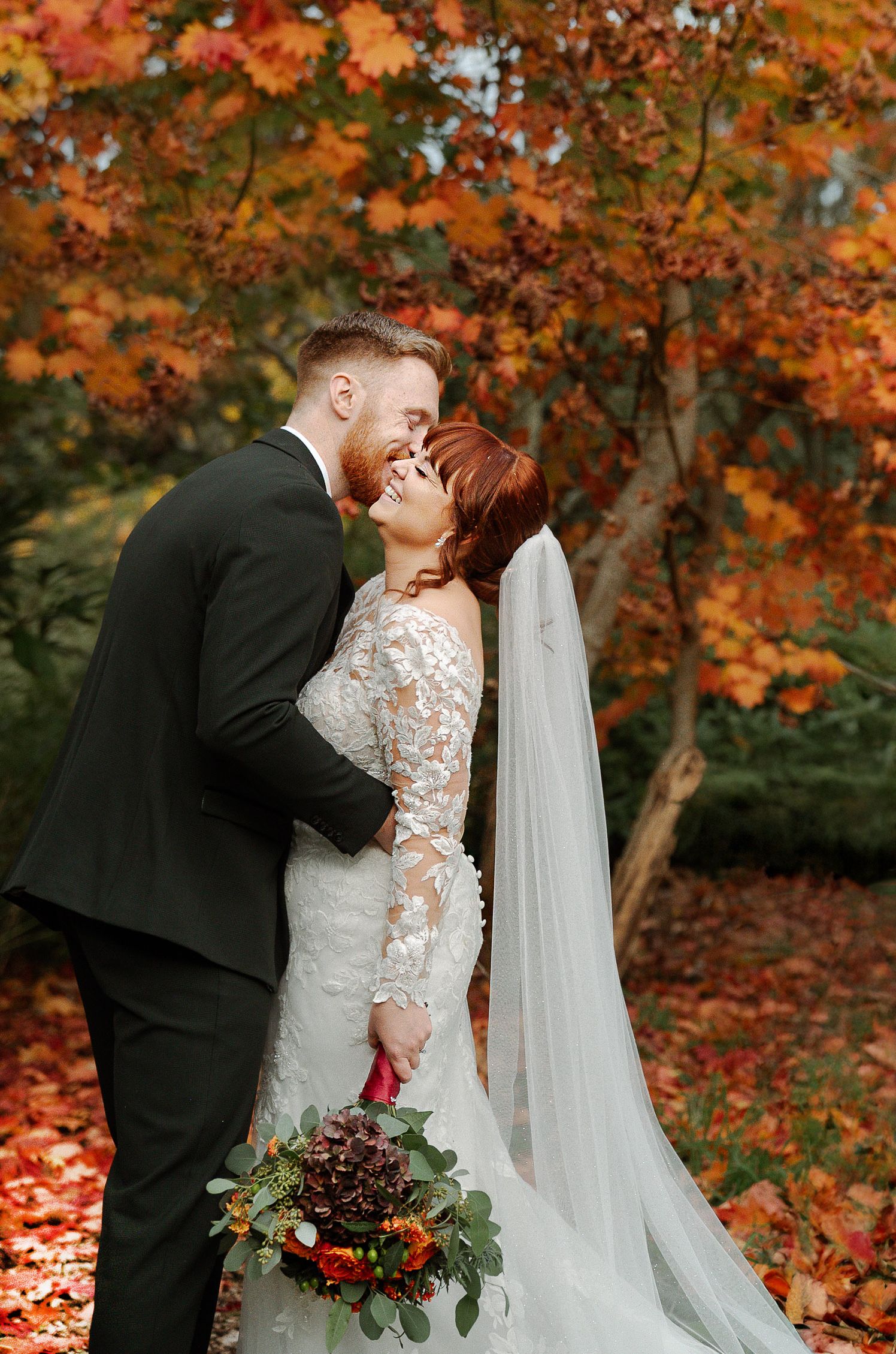 A couple shares a romantic kiss surrounded by vibrant orange autumn foliage during their fall wedding ceremony.
