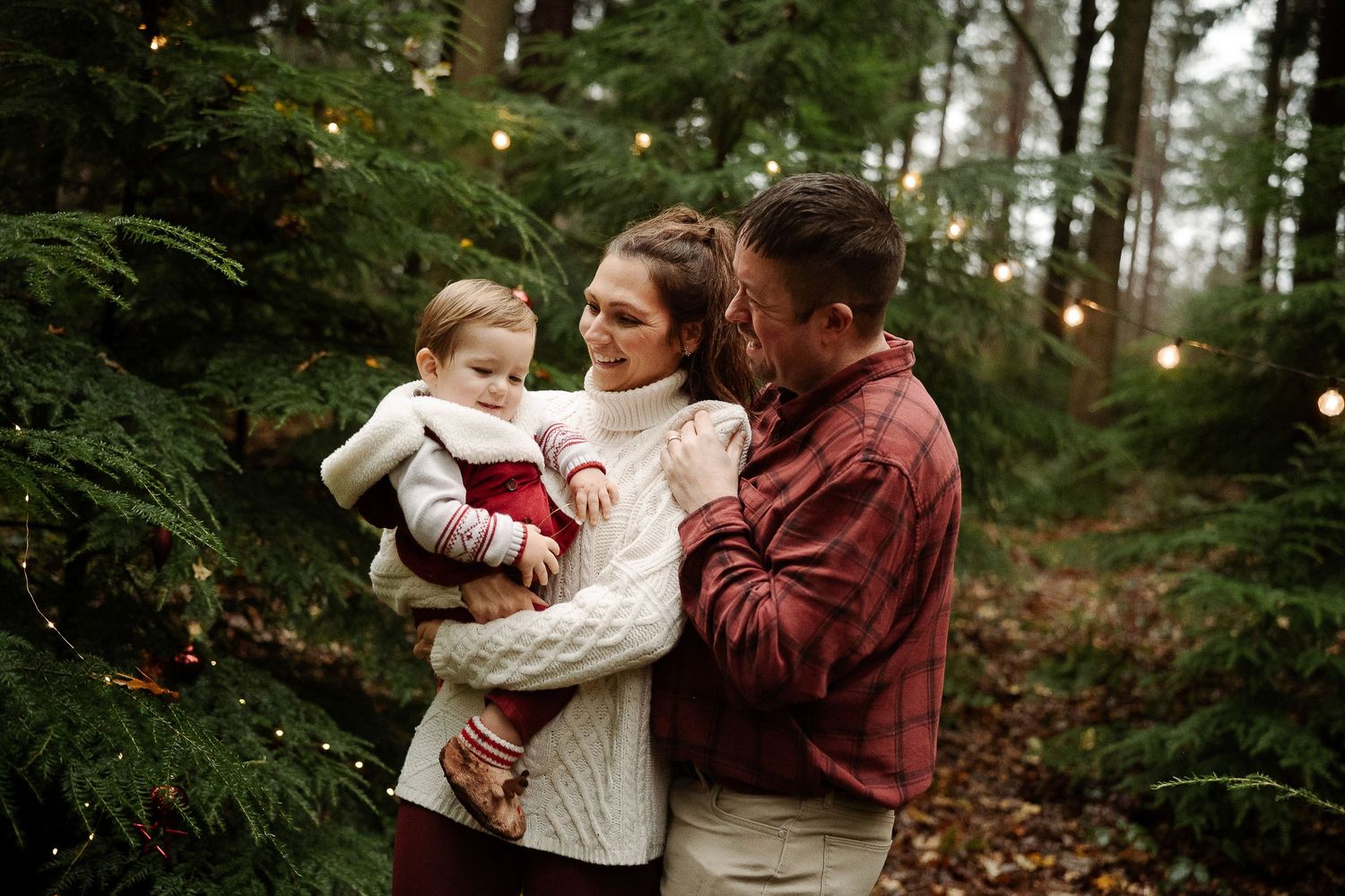 A family shares tender moments together in a forest setting with evergreen trees and soft lighting.