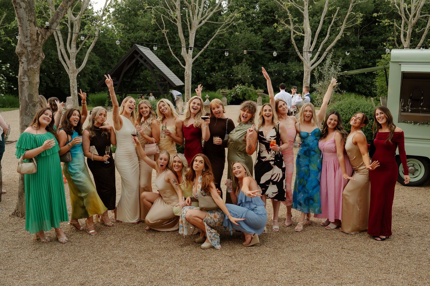 Large group of wedding guests in colorful dresses pose together outdoors in a garden setting for a group photo.