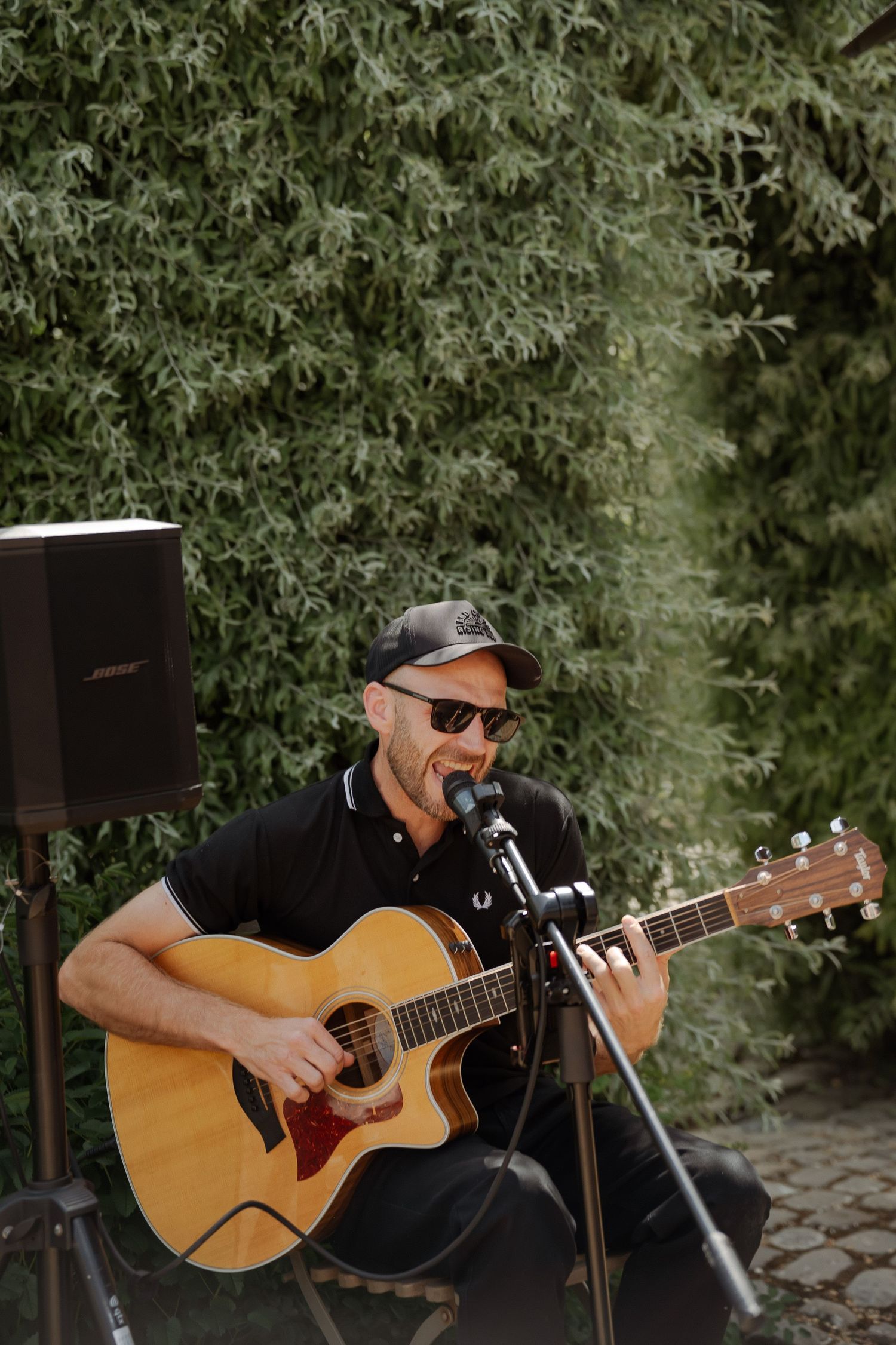 Acoustic guitarist performs outdoors among green foliage at a wedding event.