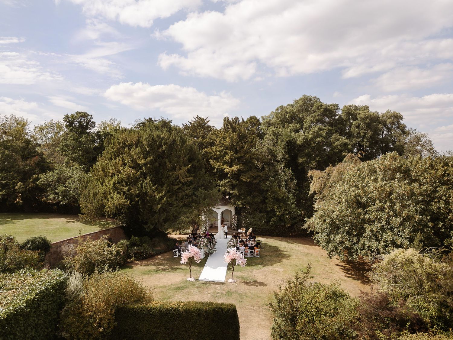 Aerial view of an outdoor wedding ceremony in a lush garden surrounded by tall trees under a partly cloudy sky.