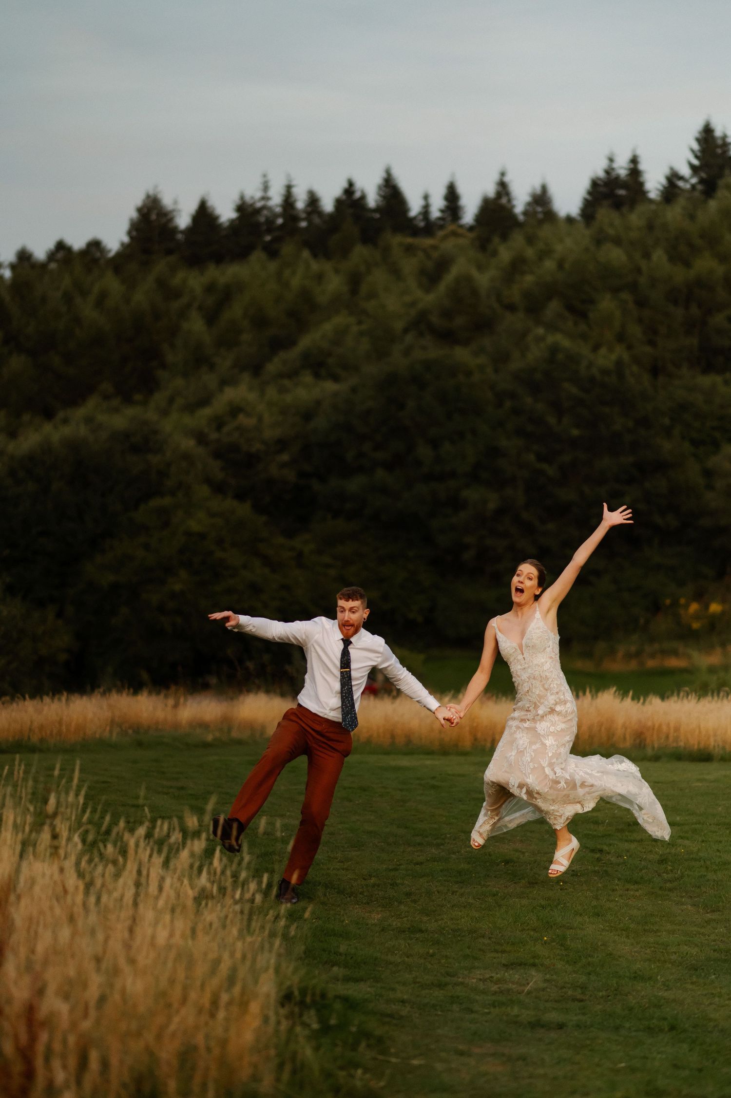 A couple joyfully leaps into the air in a meadow with evergreen trees silhouetted against an overcast sky.