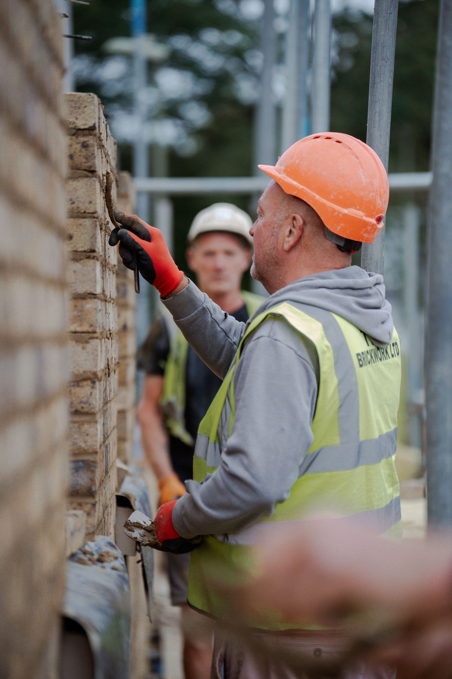 Construction worker in safety vest and orange hard hat applies mortar to brick wall at building site.