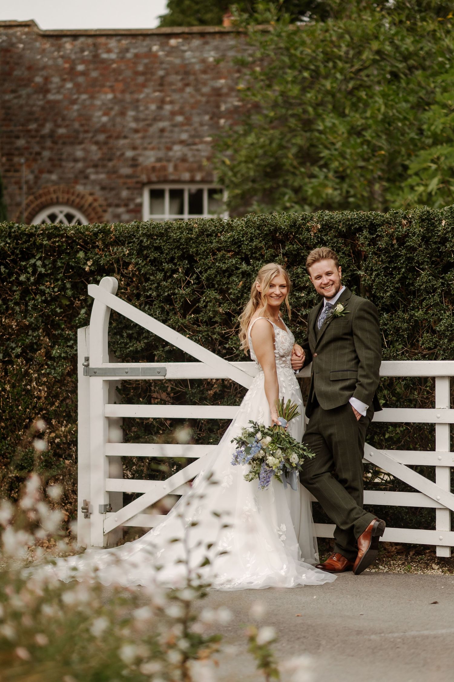 Bride and groom posing by a white wooden gate at a countryside wedding venue surrounded by lush greenery.
