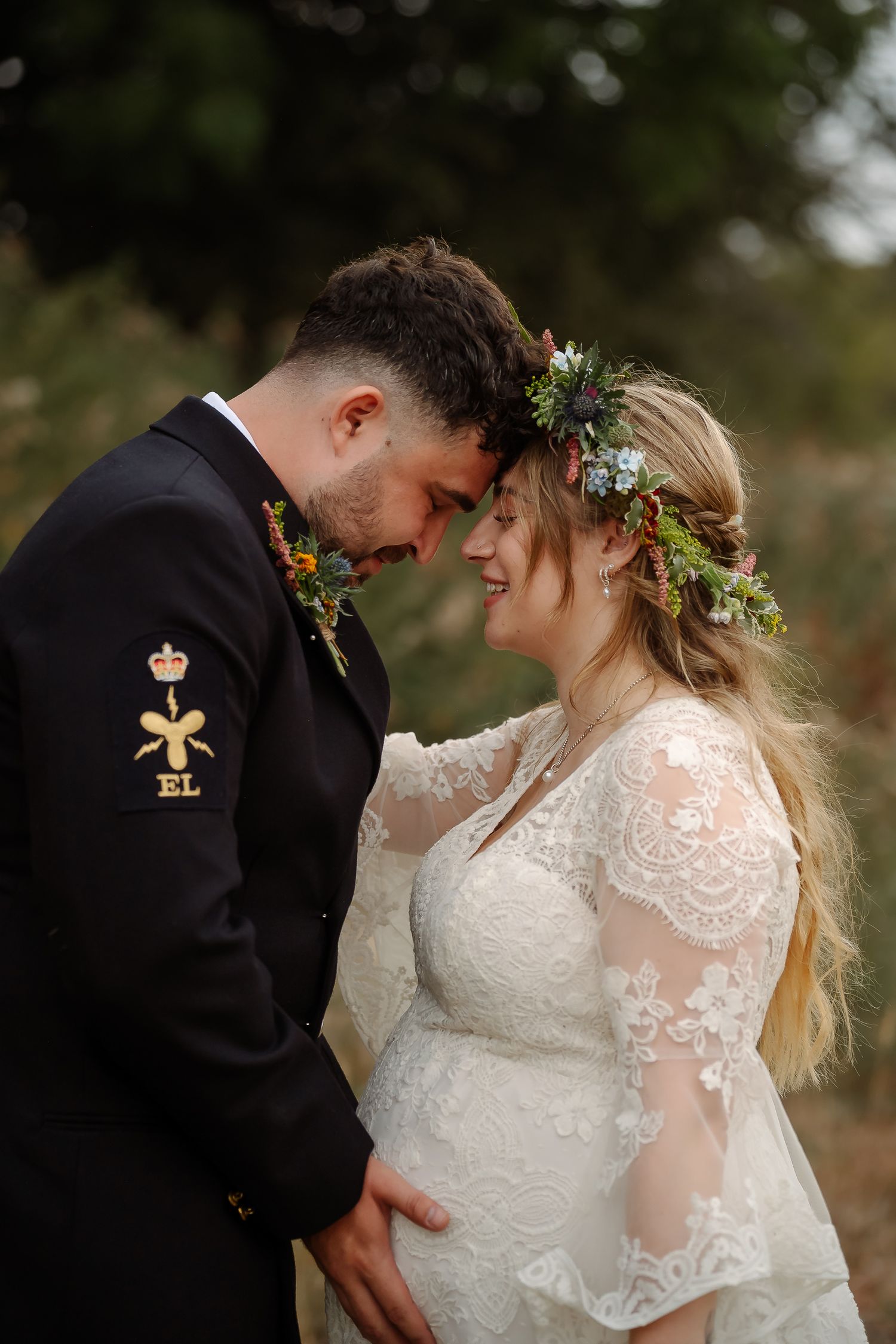 A bride in lace dress with floral crown and groom in military uniform share an intimate forehead touch outdoors.