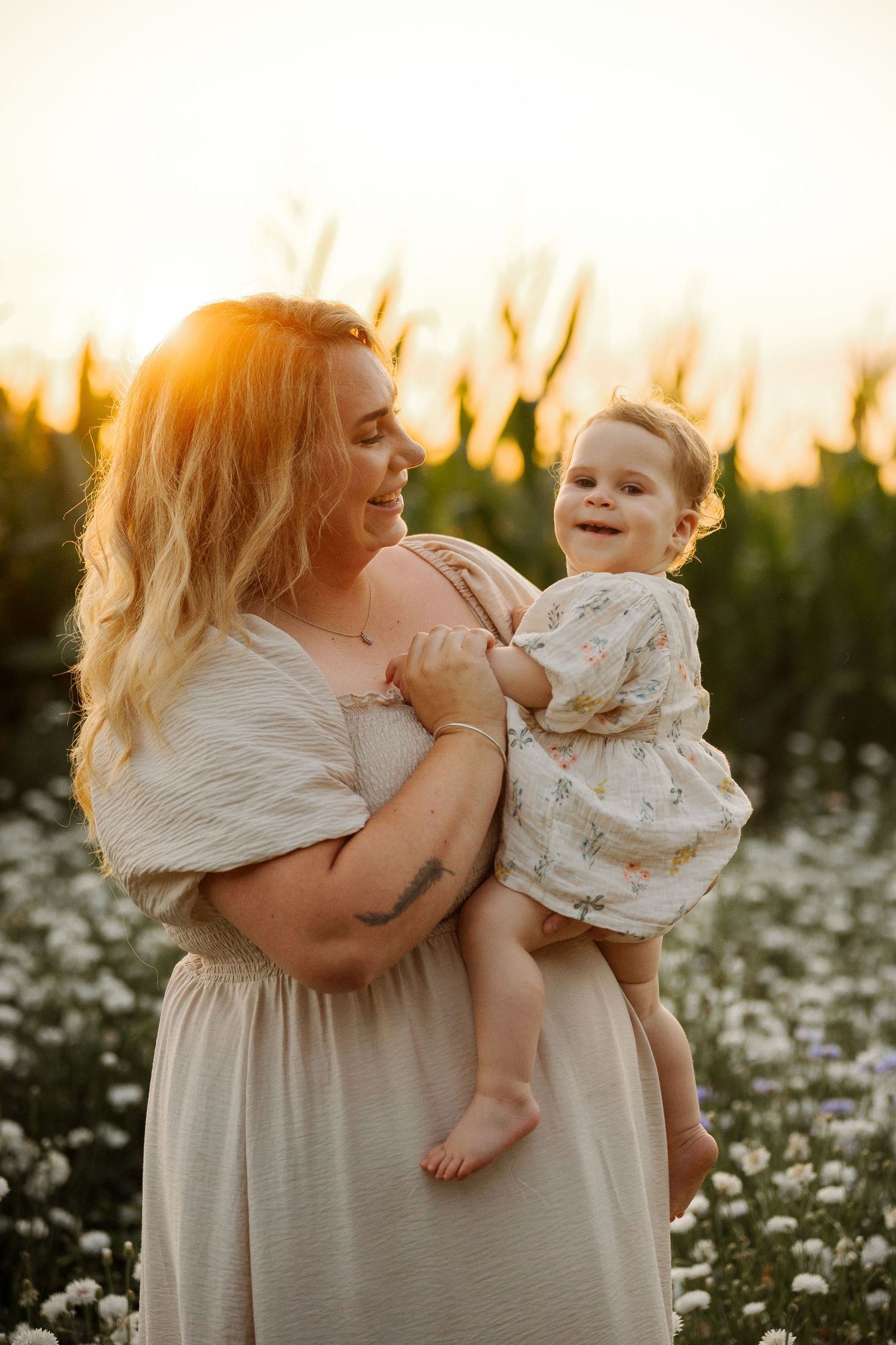 Mother holding smiling toddler in floral dress in a wildflower field at golden sunset.