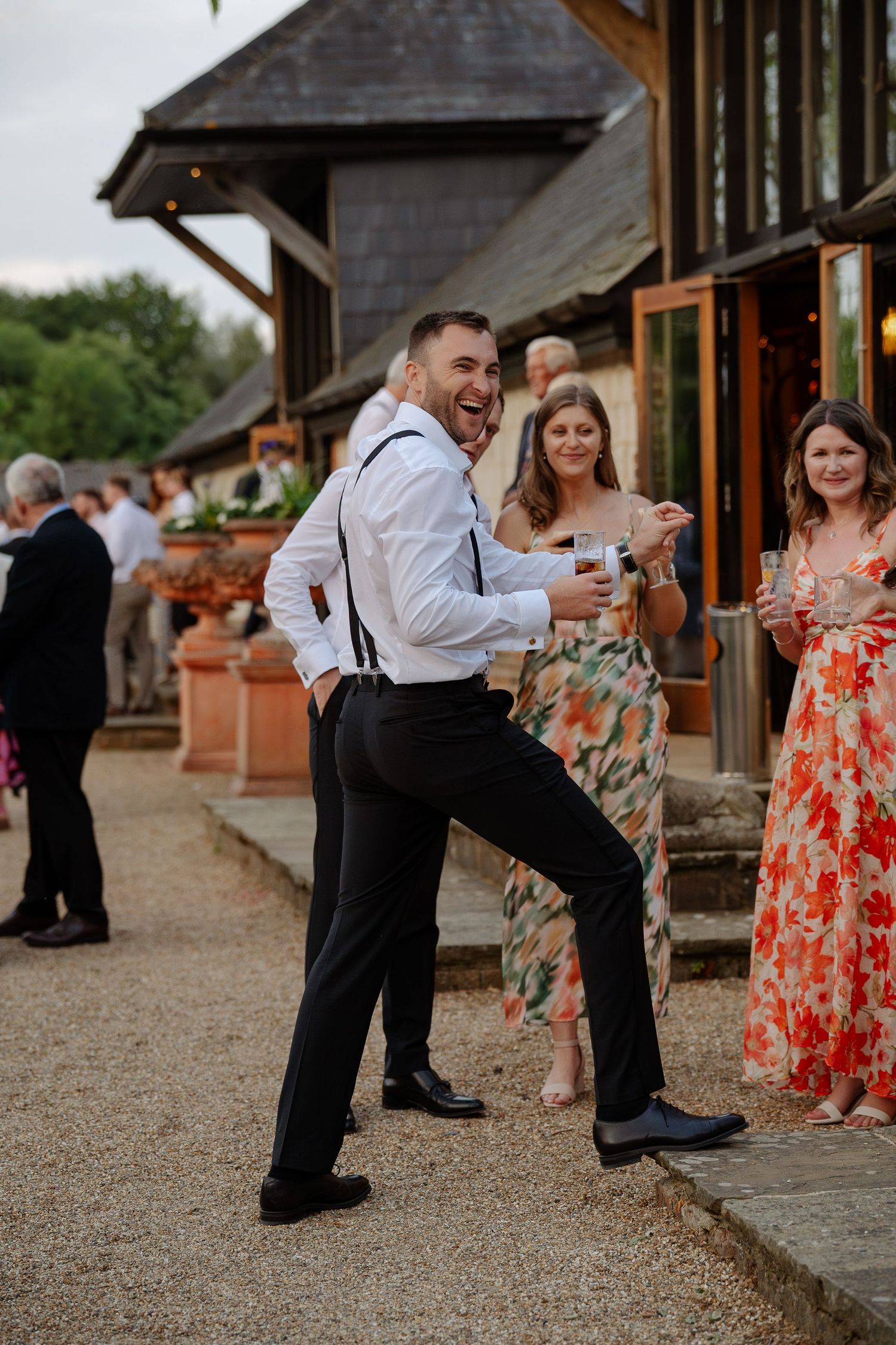 Wedding guests in colorful attire gather outside rustic venue while man in suspenders strikes playful pose.