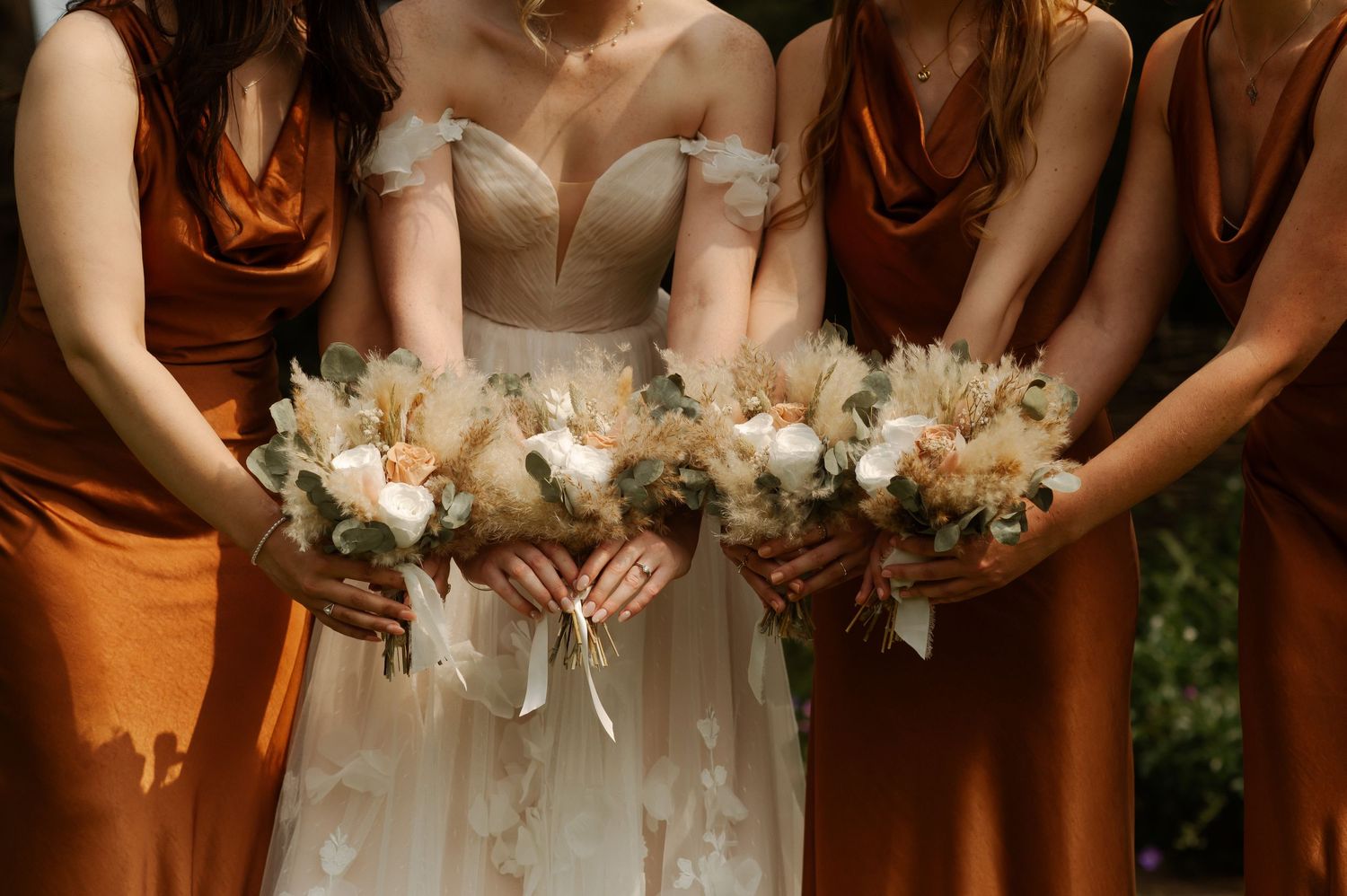 Floral bouquets held together by bridesmaids in rust orange dresses and a bride in white during an outdoor wedding.