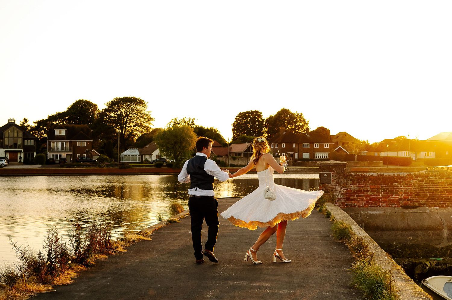 A couple dances together on a waterfront path during golden hour sunset with waterfront homes in the background.