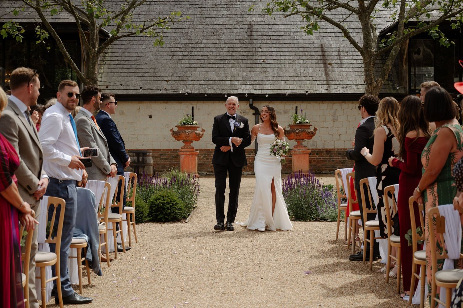 Outdoor wedding ceremony at a rustic venue with guests seated in rows on wooden chairs during a summer day.