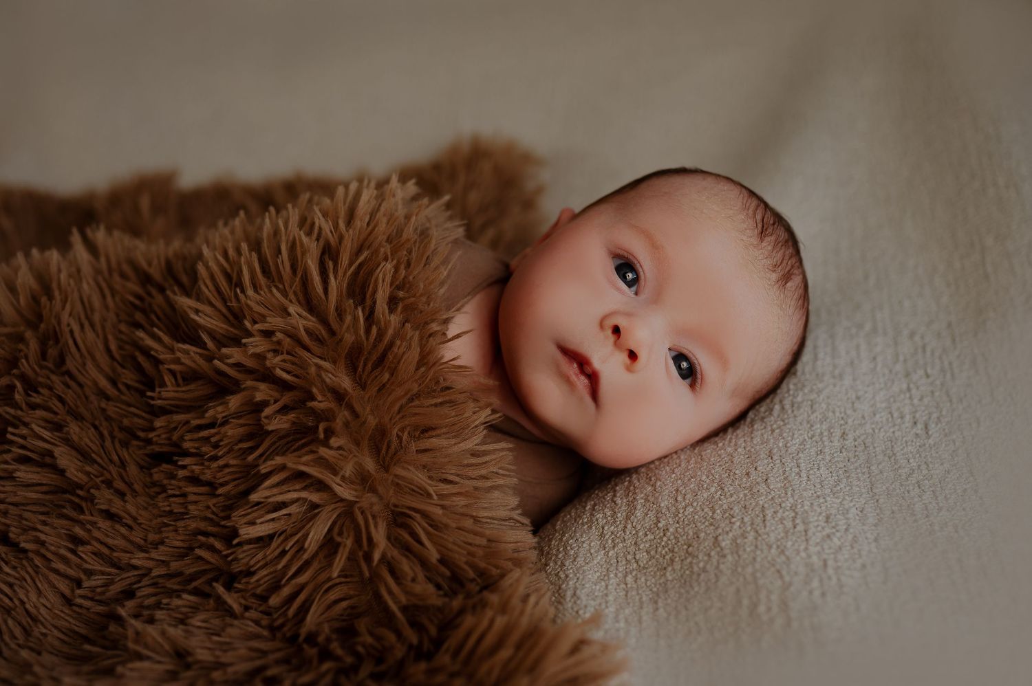 A baby wrapped in a soft brown furry blanket lies on a light colored surface.