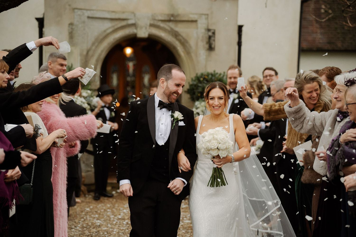 Wedding couple exits church ceremony while guests throw white confetti in a joyful celebration moment.