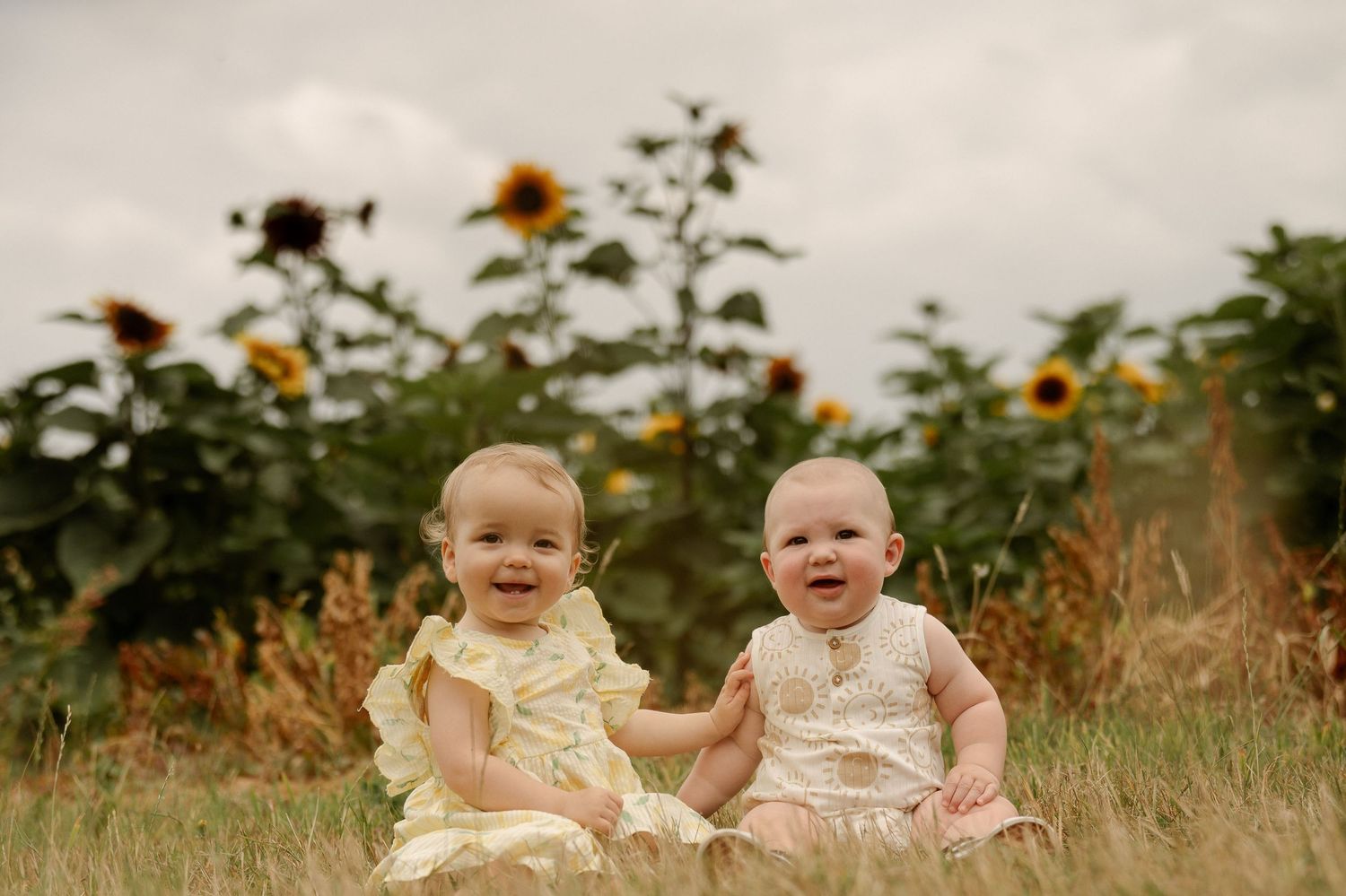 Two smiling babies in yellow dresses sitting in a sunflower field on a cloudy day.