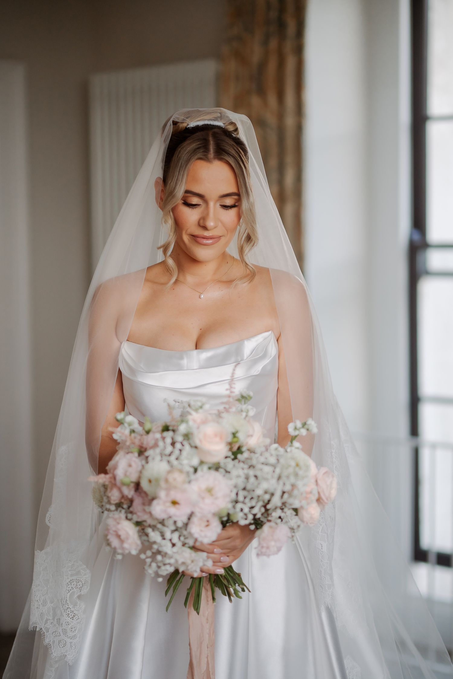 Bride in strapless white gown and veil holds pink and white floral bouquet, smiling softly indoors.