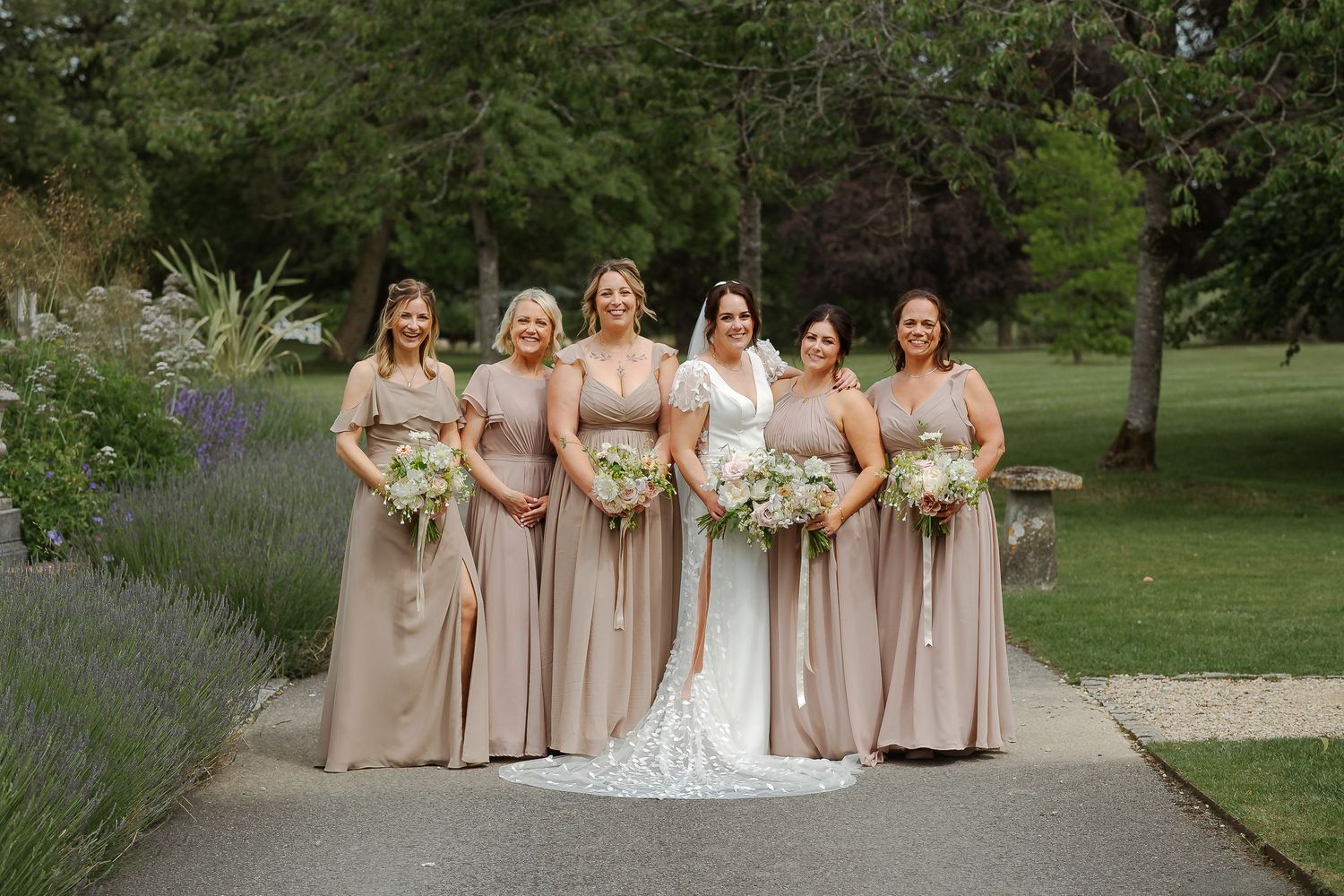 Bride in white gown poses with five bridesmaids in champagne dresses holding bouquets in a lush garden setting.