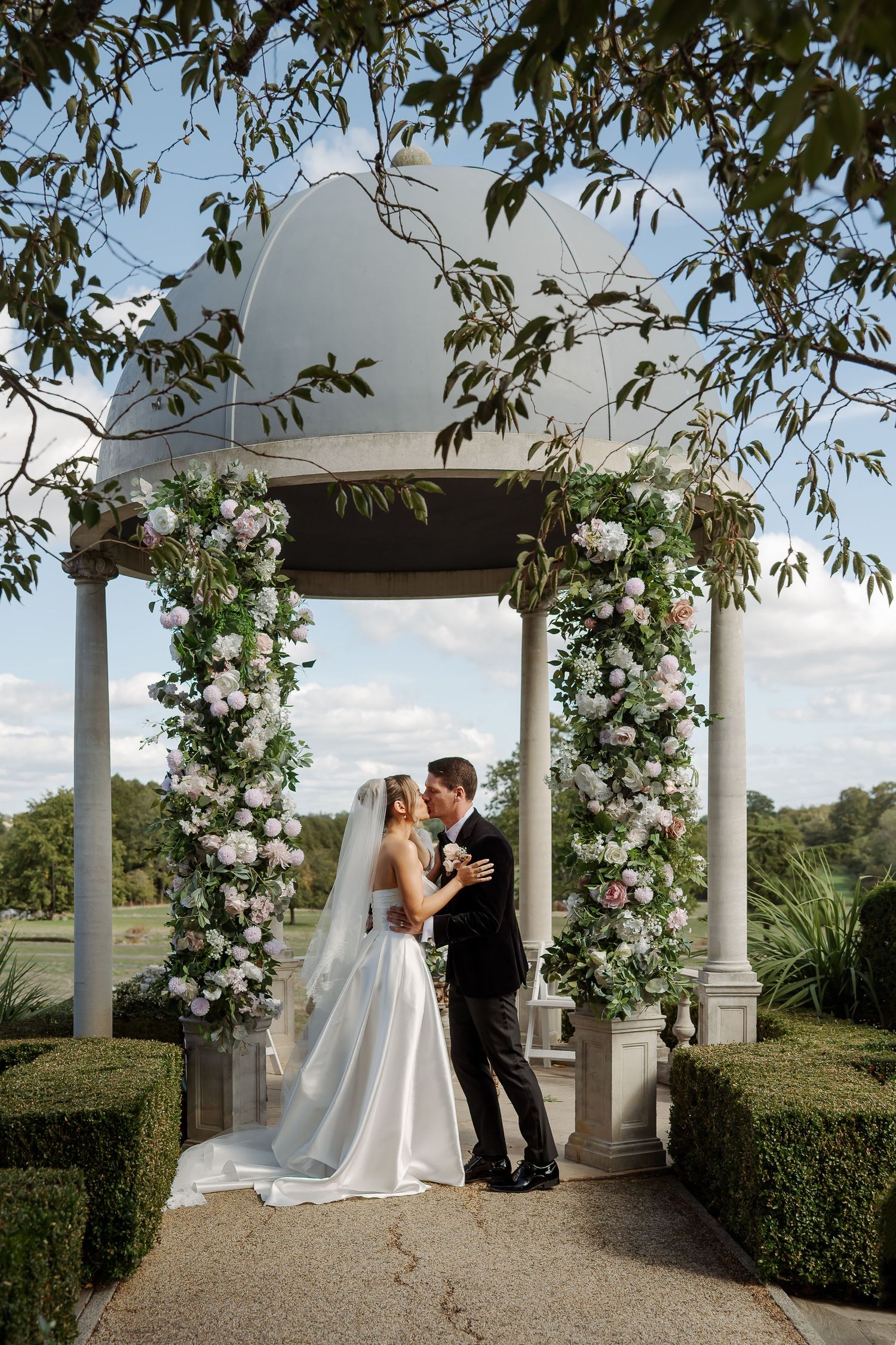 Bride and groom share a kiss under a white gazebo adorned with pink and white floral arrangements.