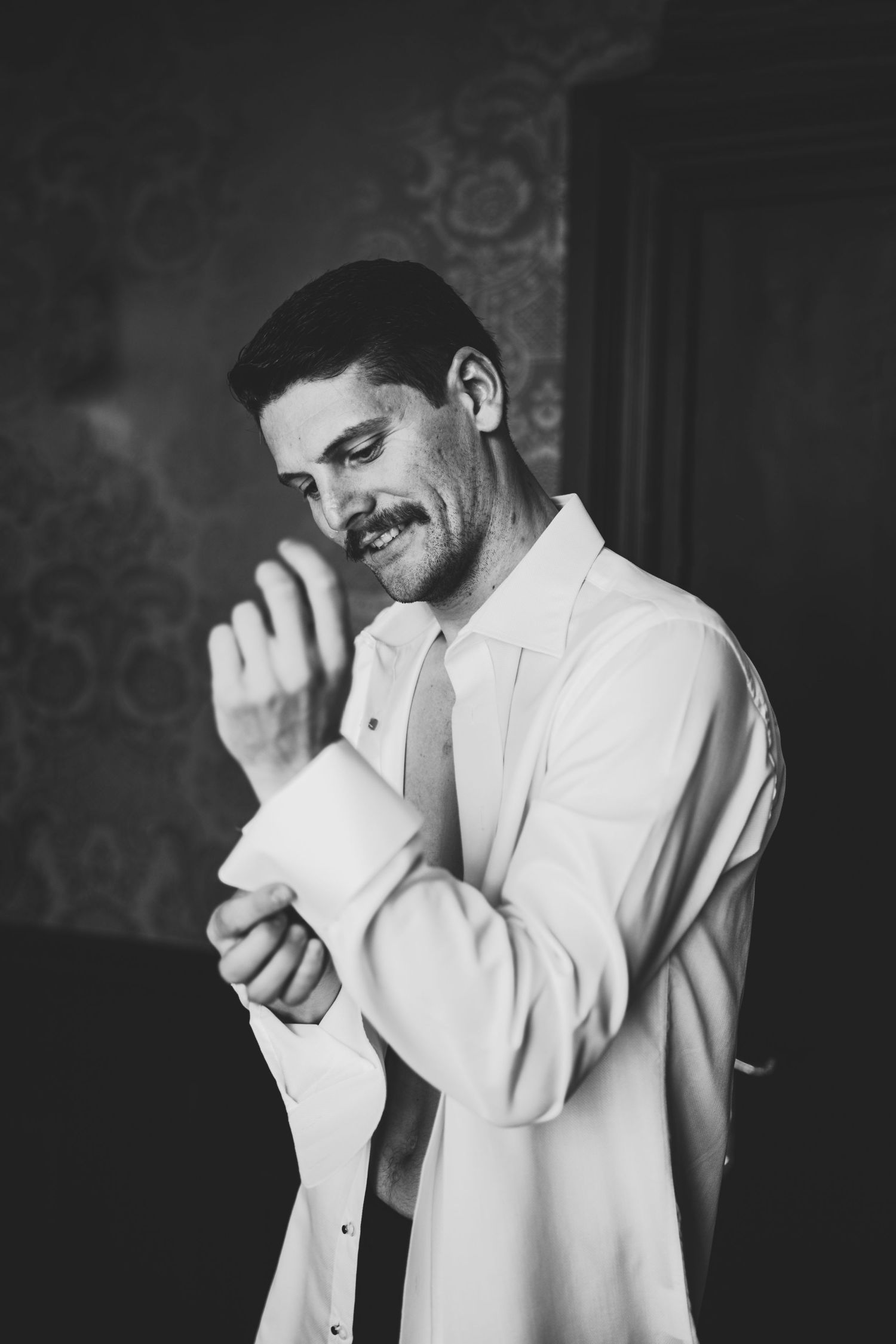 Black and white photo of a groom buttoning his shirt cuff while getting ready for his wedding day.