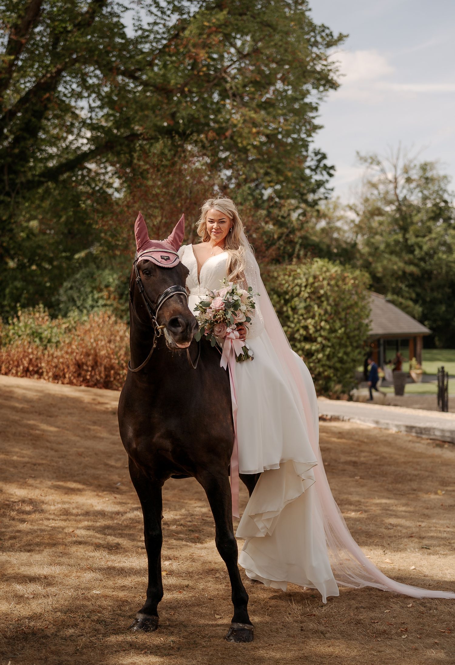 Bride in white gown holding bouquet stands beside dark horse with purple ear bonnet in garden setting.