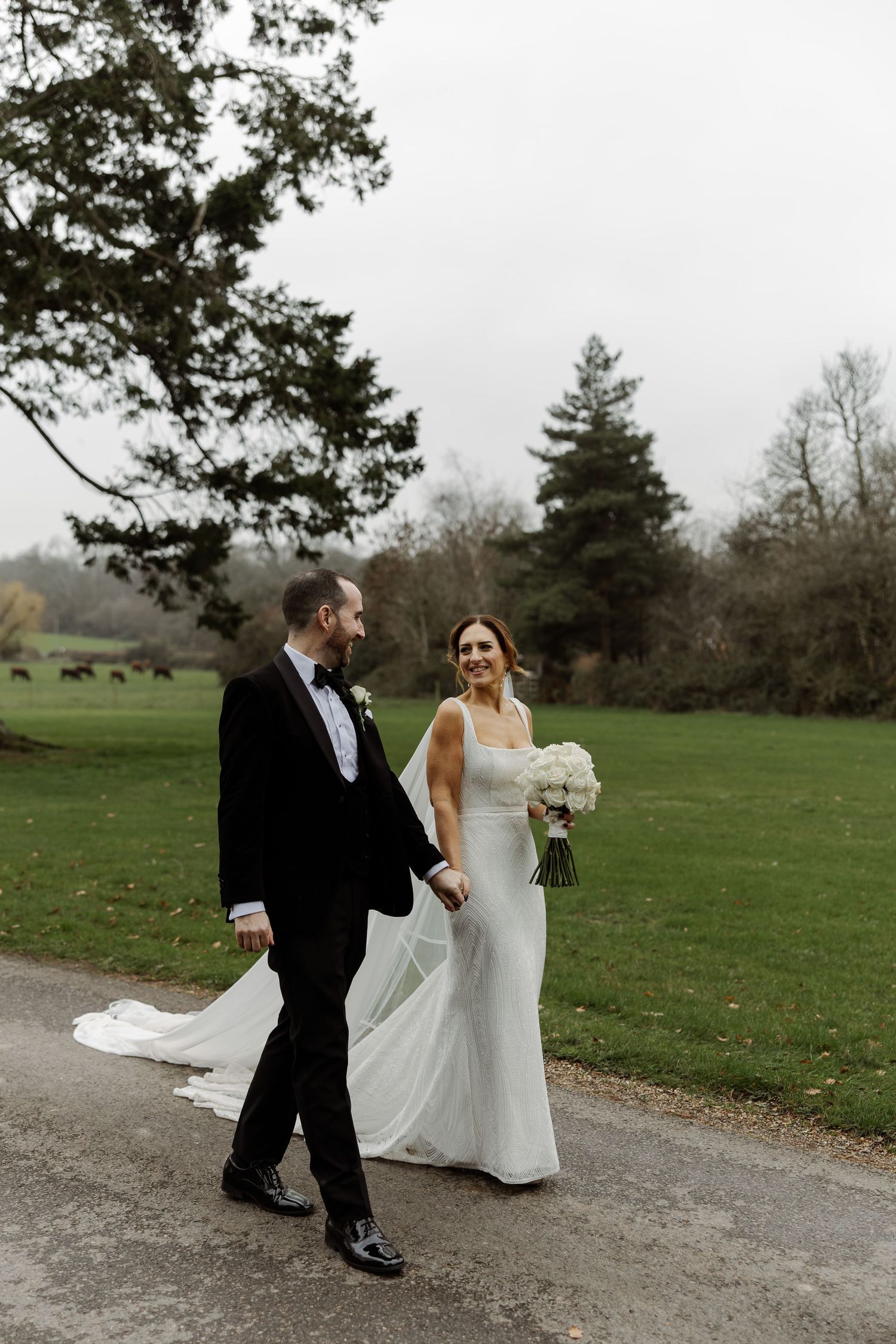 A couple in wedding attire walks and laughs together on a gravel path surrounded by green fields and trees.