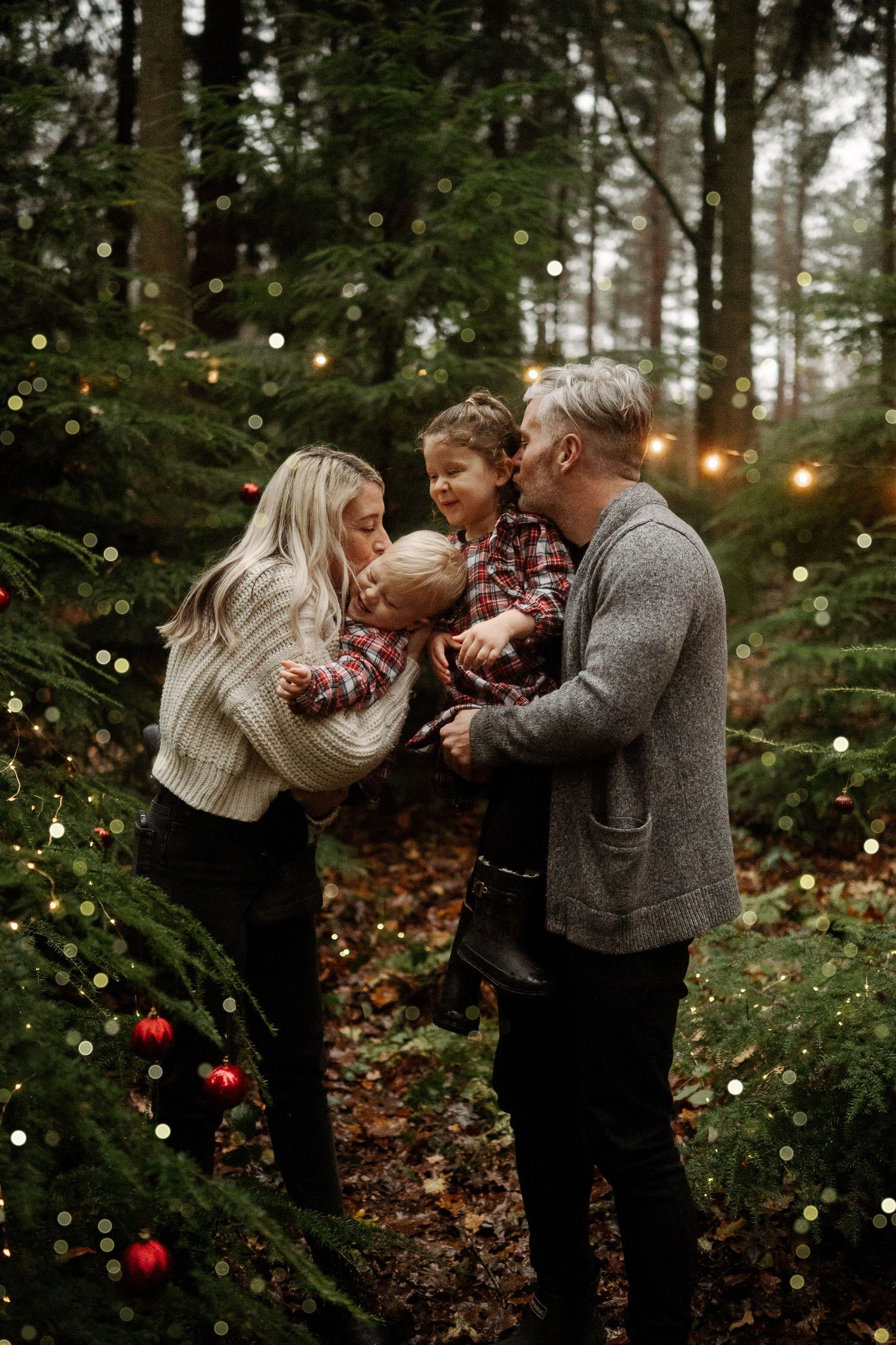 Family of three sharing kisses surrounded by Christmas trees with twinkling lights in a festive forest setting.