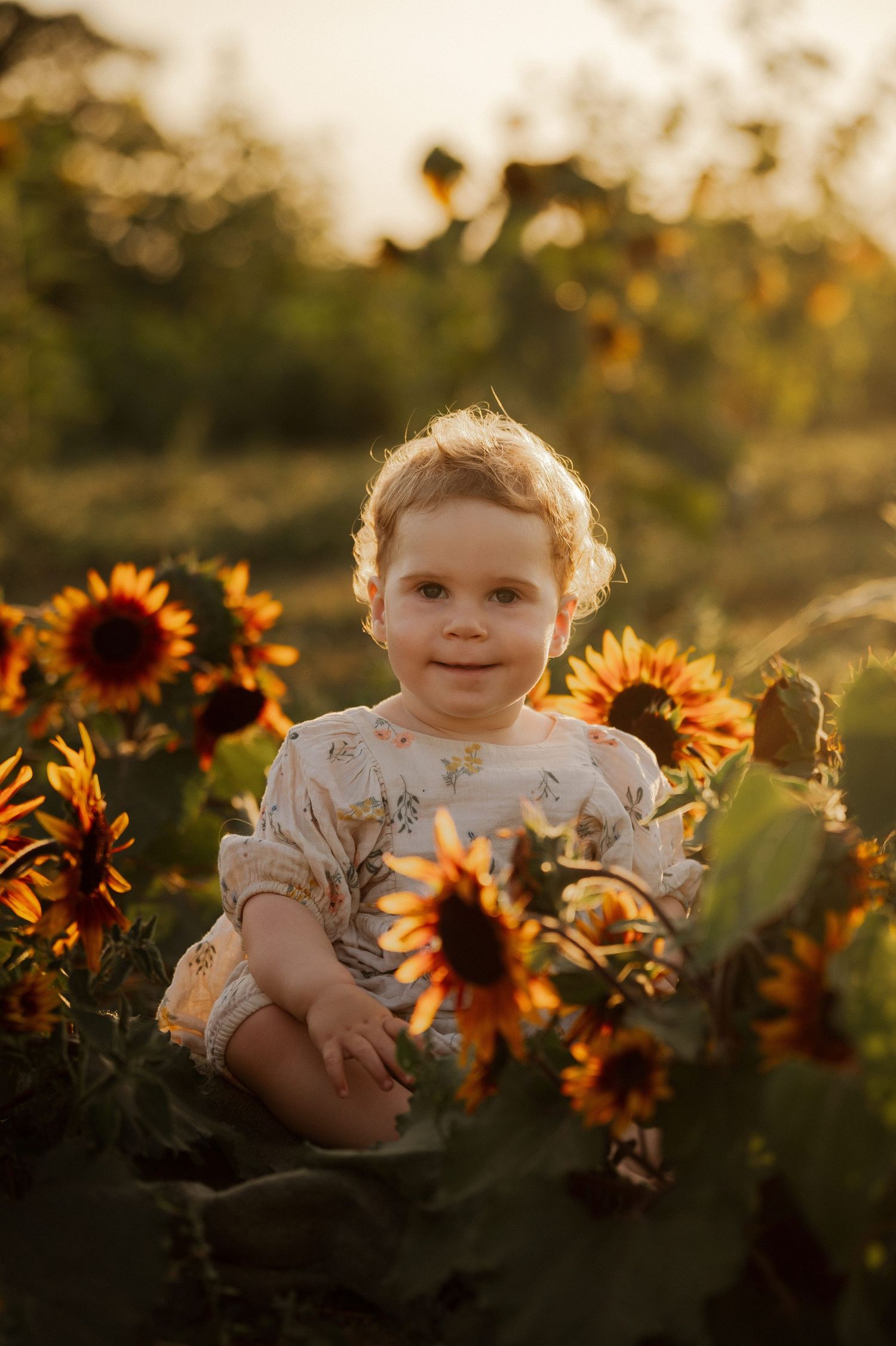 A smiling toddler sits among vibrant sunflowers in a golden sunset field, wearing a floral romper.