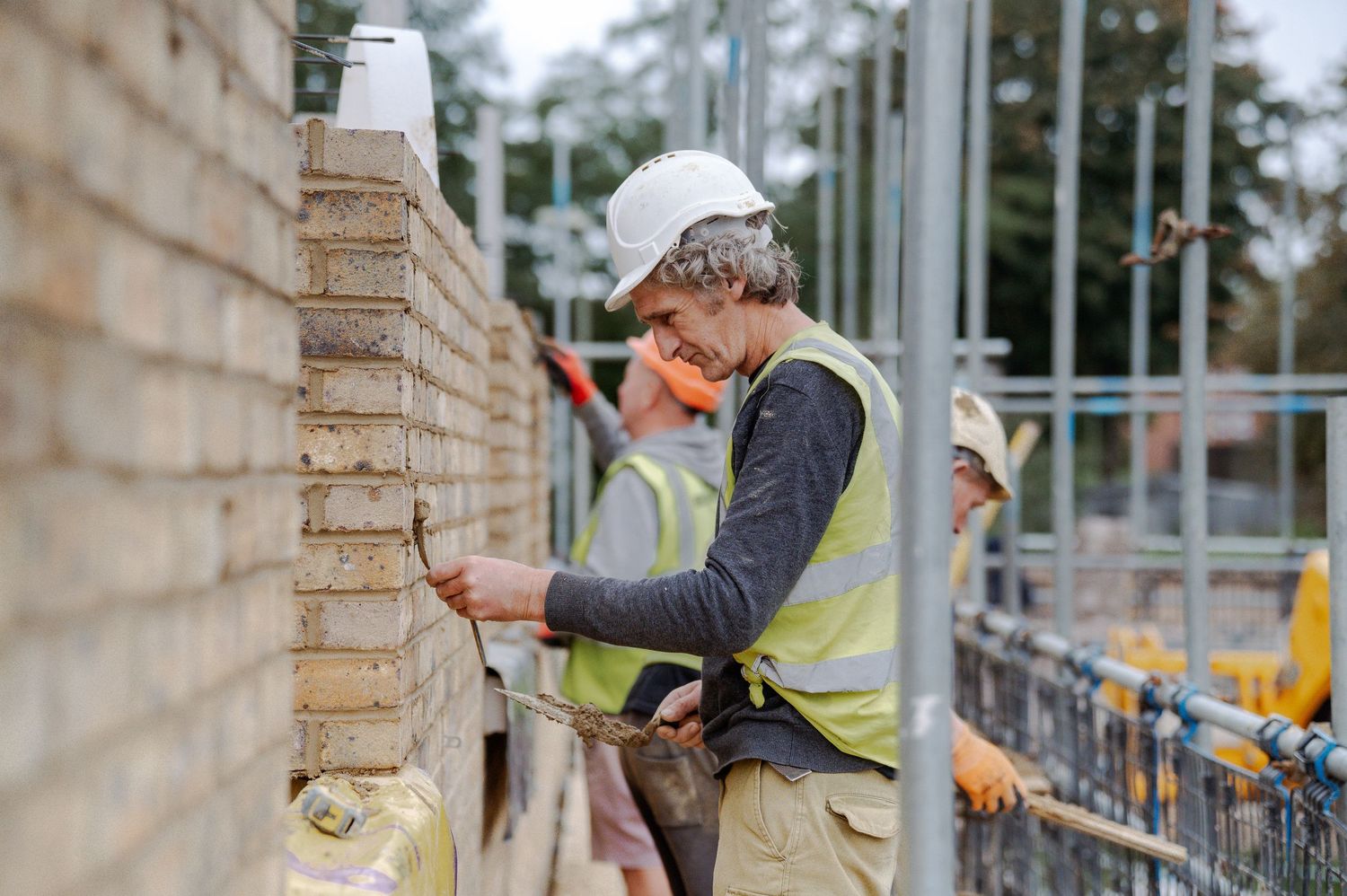 A person in safety gear uses a drill to secure metal railing posts to a brick wall.