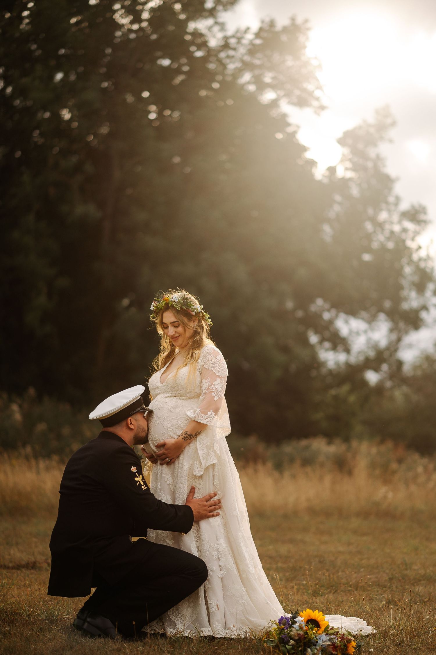 A military groom kneels before his pregnant bride in a white lace dress and floral crown, bathed in golden sunlight outdoors.