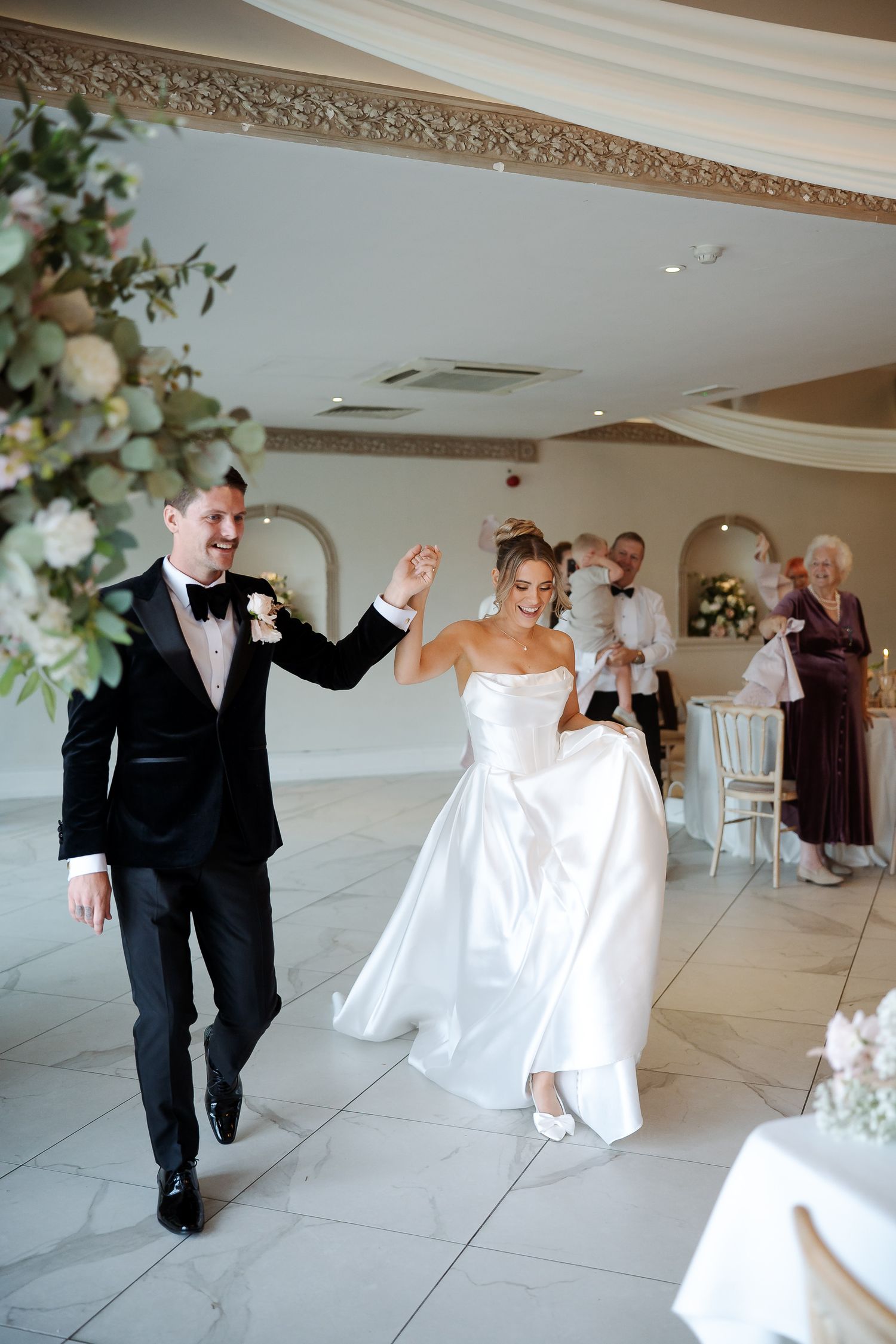 Bride and groom make grand entrance into elegant wedding reception hall, bride lifting dress, guests applauding in background.